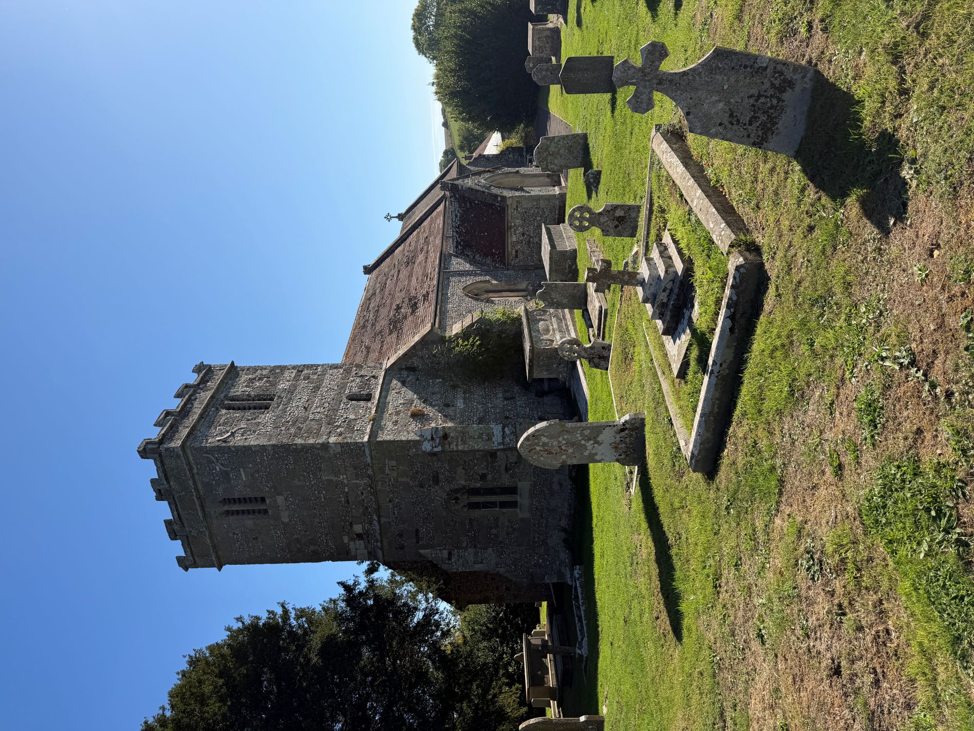 A small grey stone and flint church with a battlemented square tower and a red tiled roof, seen across a grassy churchyard with lichen-covered headstones, crosses and a low kerbed grave, under a clear blue sky.