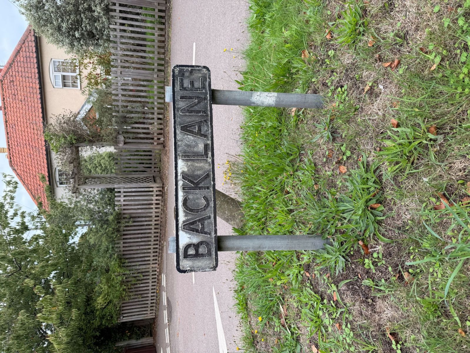 A black and white street sign reading Back Lane on two posts in a grassy verge, with a road, a wooden fence and a house with a red tiled roof behind.