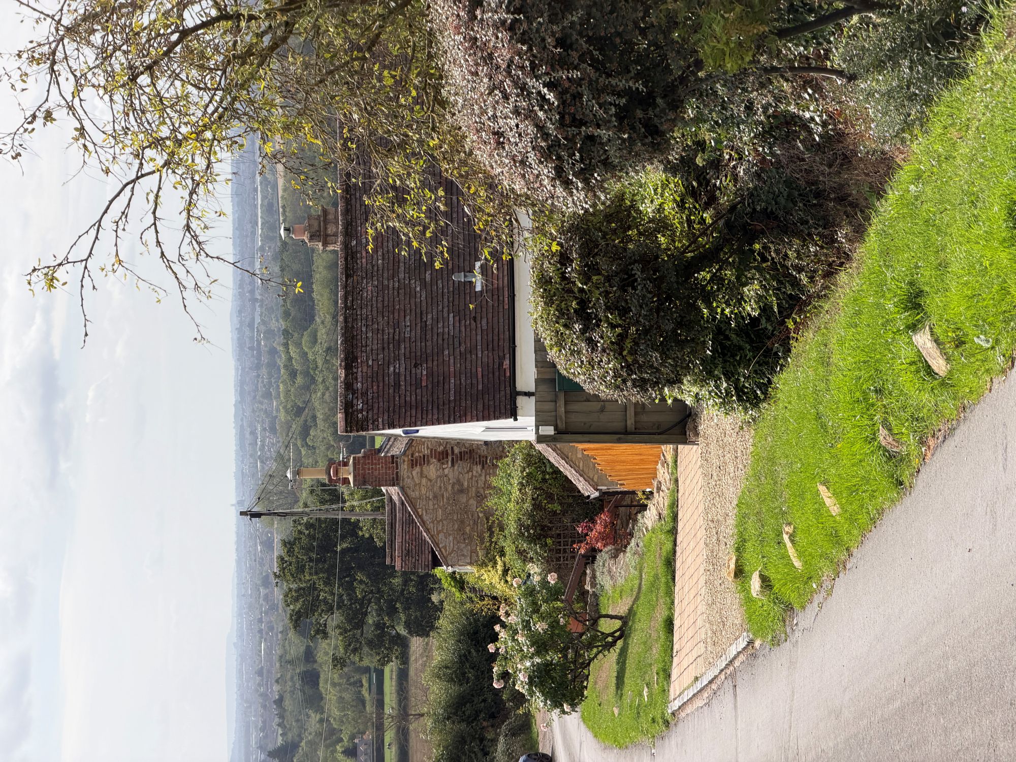 A narrow road curving downhill past a white-painted cottage and a stone cottage with tiled roofs and brick chimneys, with a wide hazy view over fields and woodland in the distance.