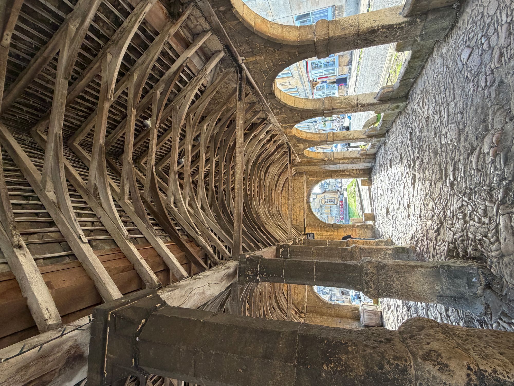 A cobbled floor beneath a vaulted timber roof supported by stone pillars and arches that open onto a street.