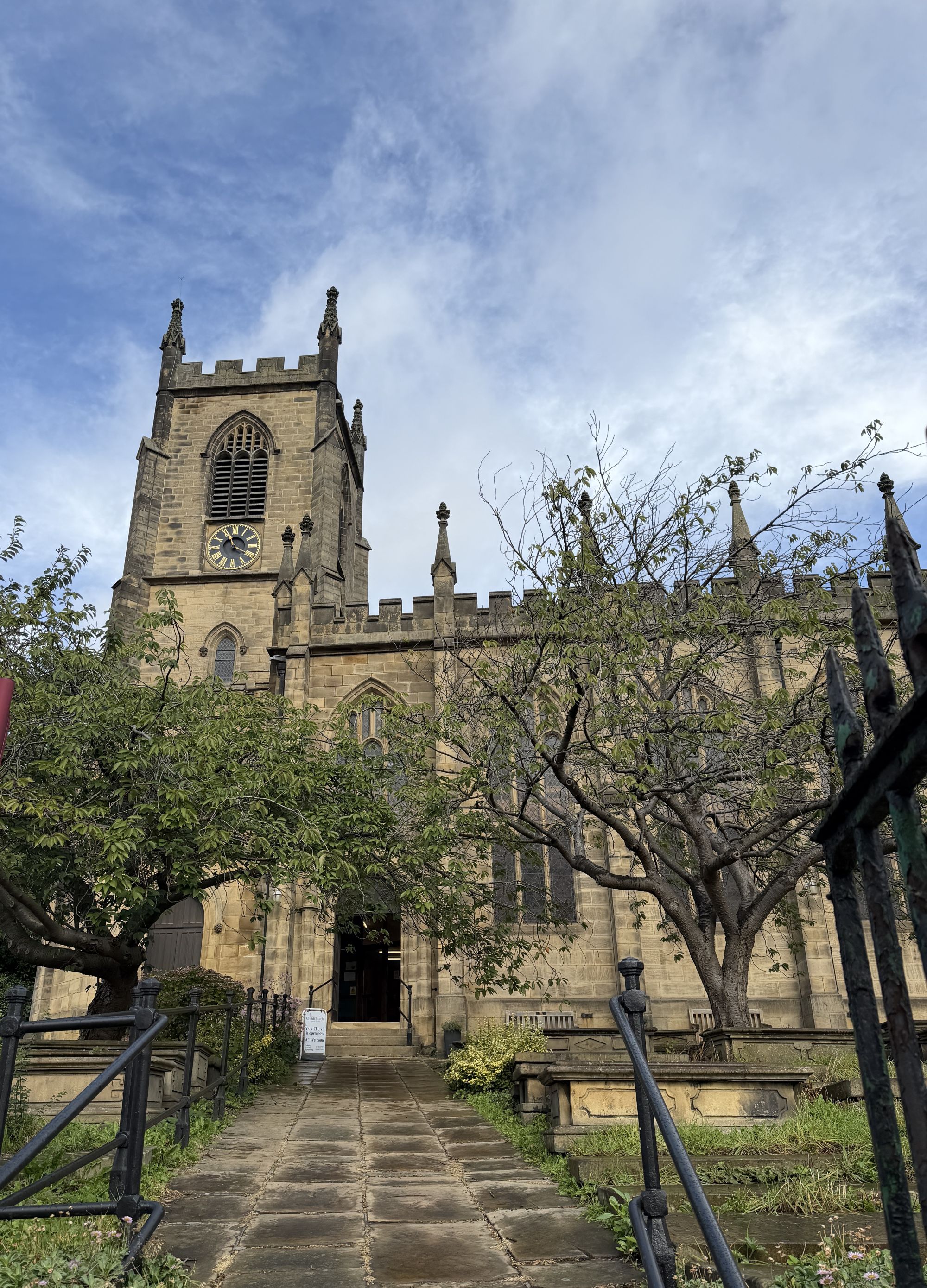 A stone church with a battlemented tower and clock, seen from a flagged path through the churchyard, with trees and iron railings in the foreground.