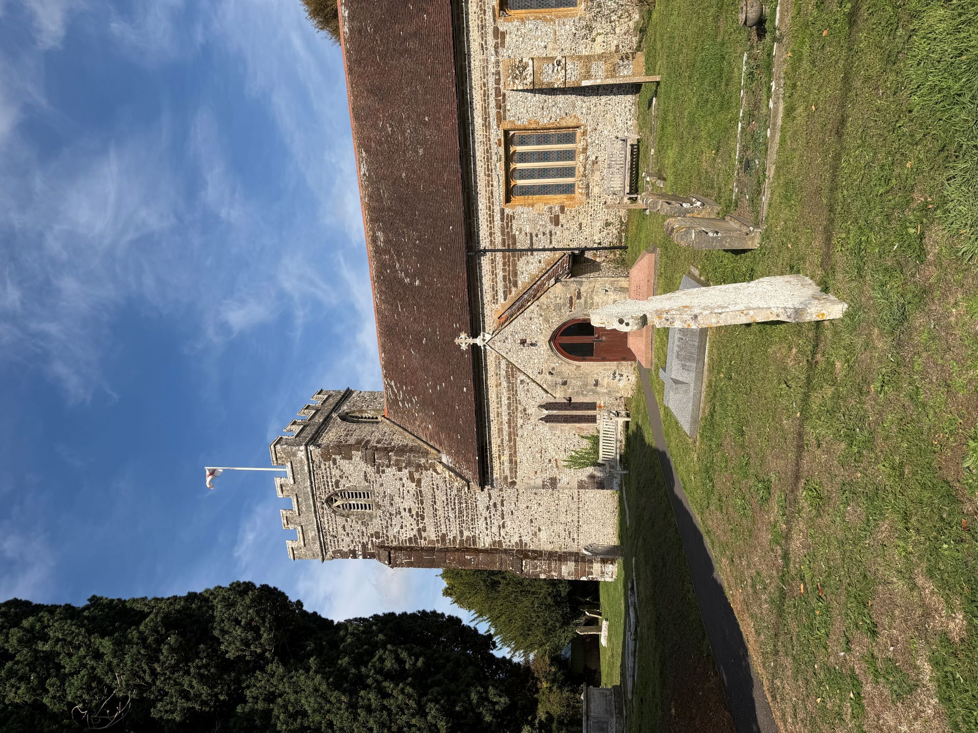 A flint and stone church with a battlemented tower flying a flag, a red tiled roof and a small gabled porch with an arched red door, seen across a churchyard with headstones and a tall dark evergreen tree, under a blue sky.