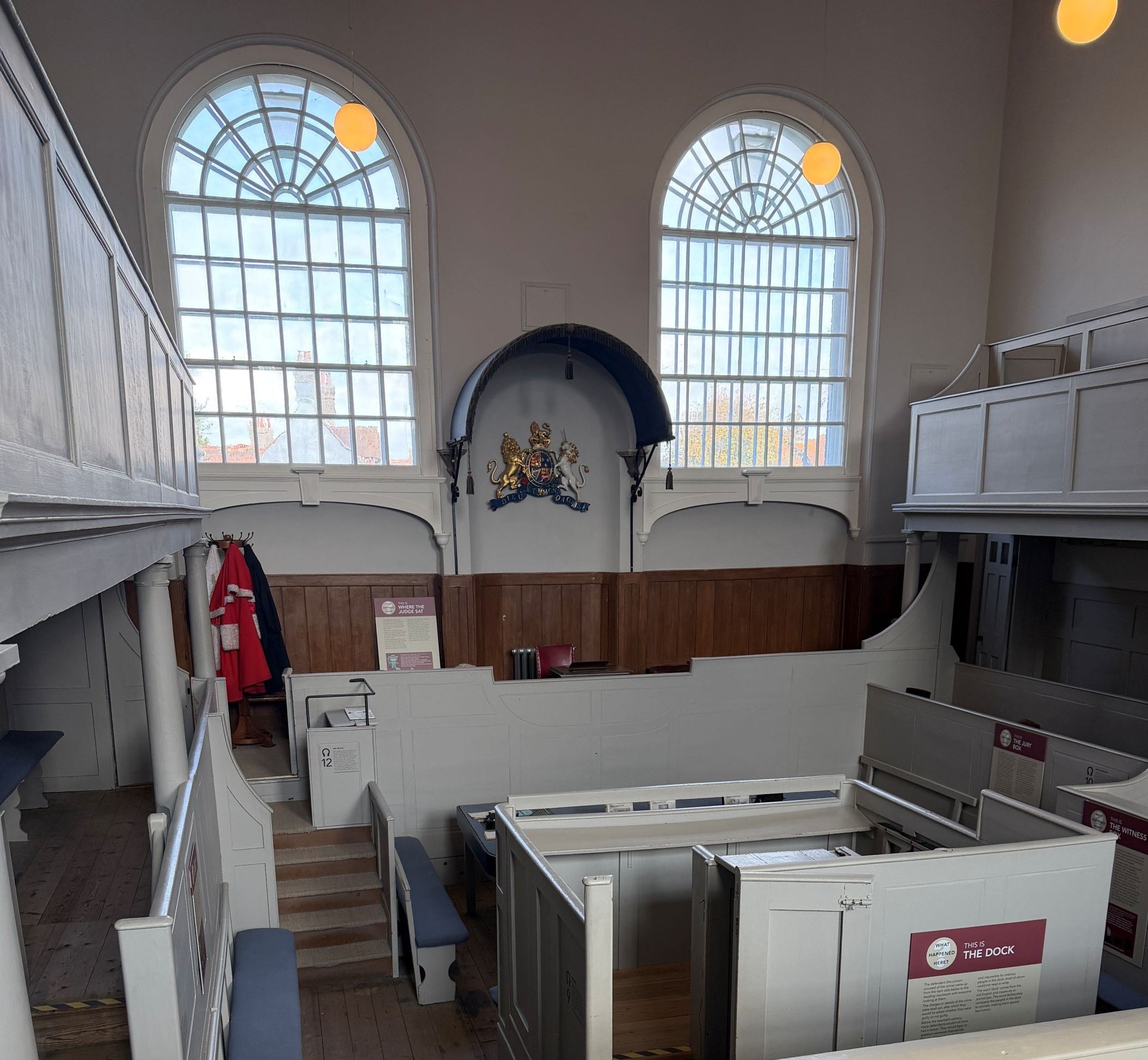 A pale painted courtroom seen from the public gallery, with tall arched windows, a royal coat of arms above the judge&rsquo;s seat, wooden panelling, and the enclosed dock in the foreground.