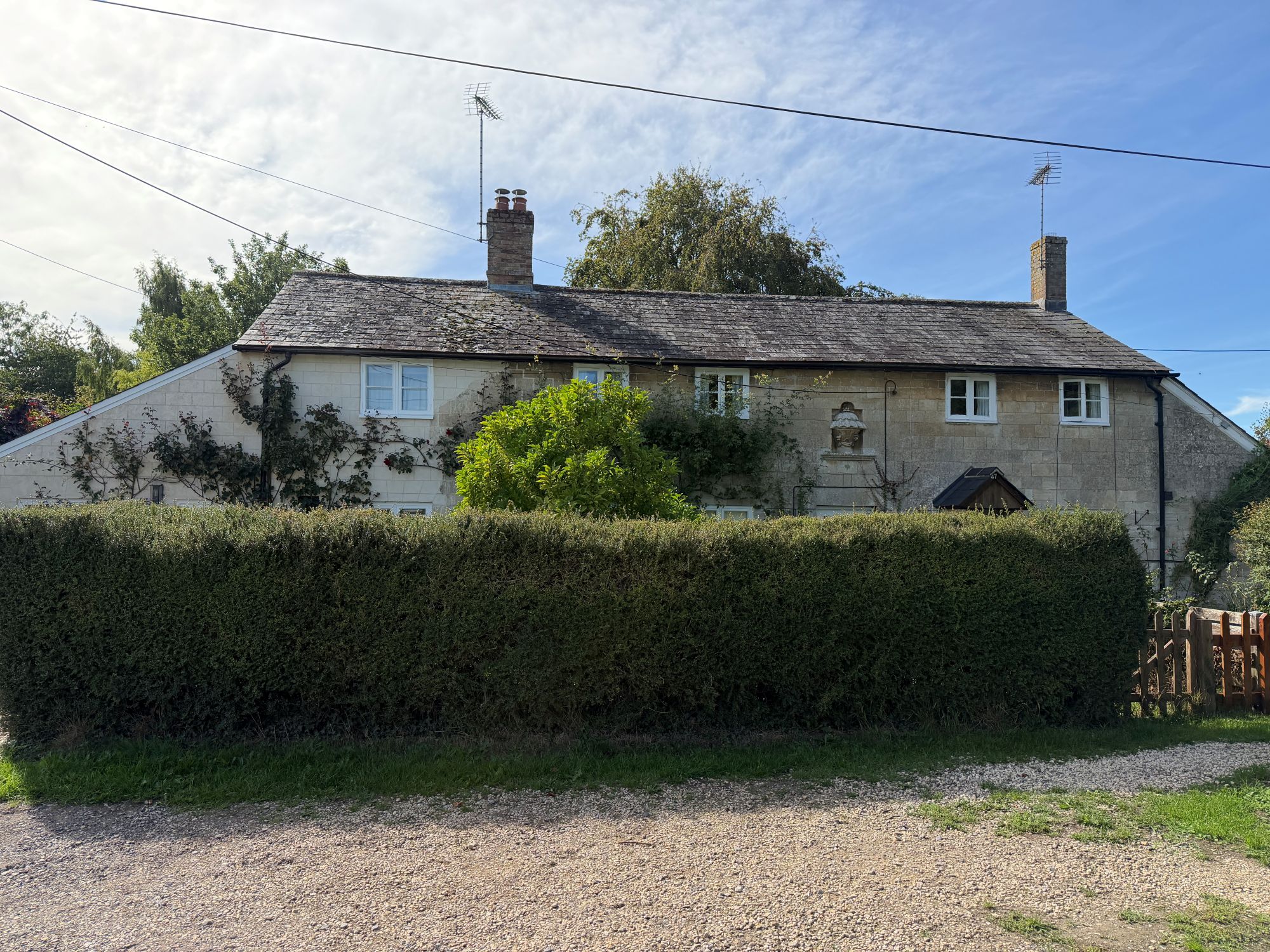 A pair of stone cottages with a slate roof and two chimneys, partly hidden behind a tall clipped hedge, with climbing roses and shrubs, under a blue sky crossed by overhead wires.
