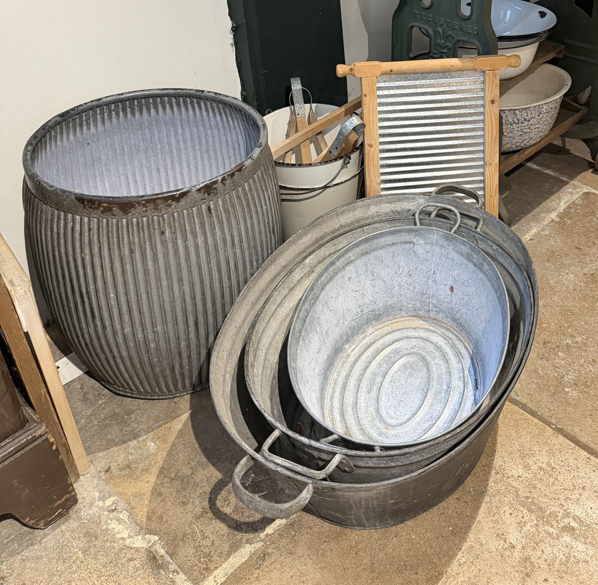 Old laundry equipment on a stone floor: a ribbed galvanised dolly tub, a stack of oval tin wash tubs with handles, a wooden-framed metal washboard, an enamel bucket holding wooden dolly pegs, and enamel bowls on a shelf behind.