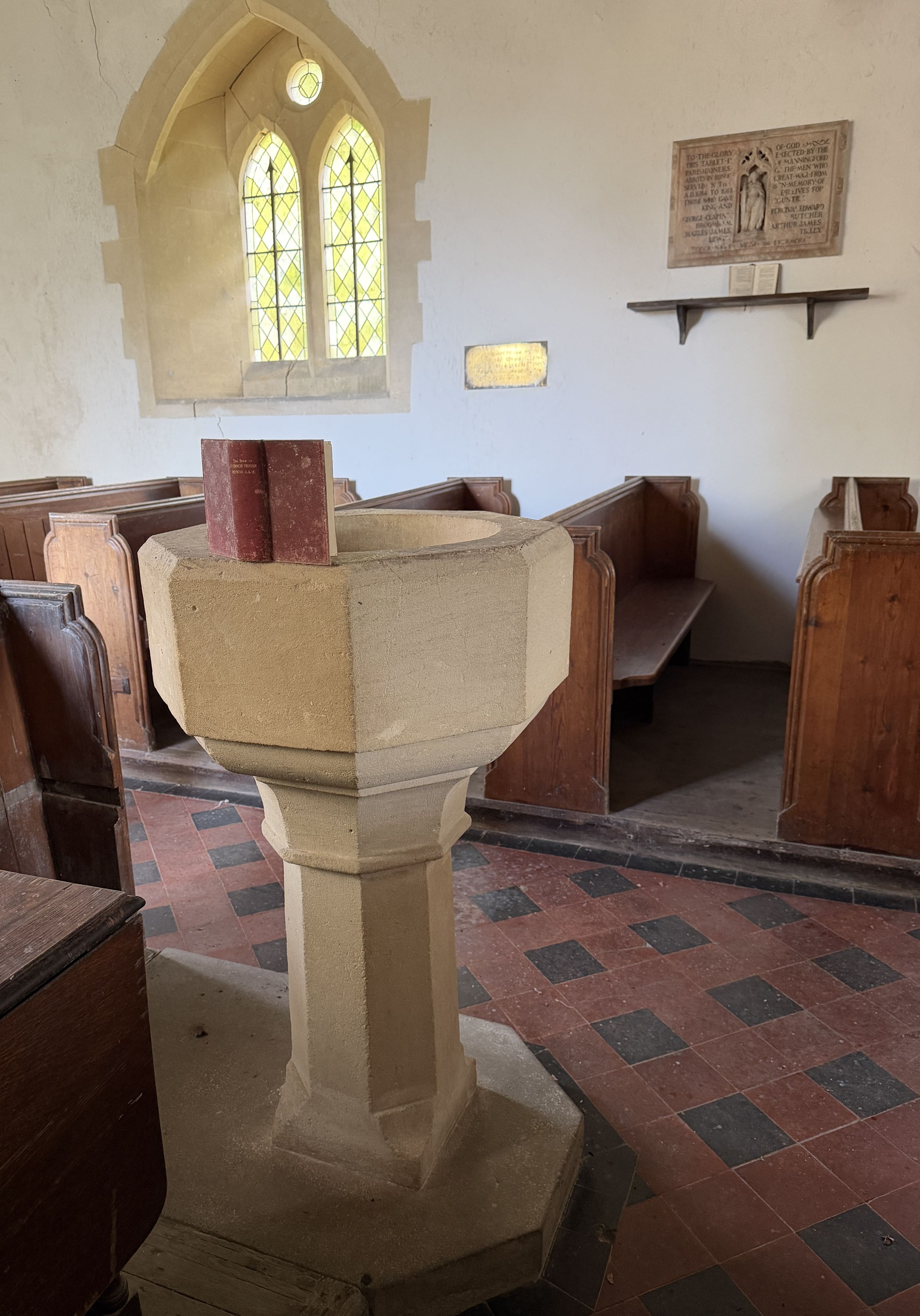 A pale stone octagonal font on a stepped base, with a red book titled "The Book of Common Prayer Hymns A&M" resting on its rim, inside a church with wooden pews, a tiled floor and a stained glass window behind.