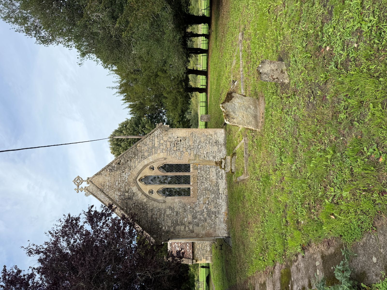 A small stone church seen from the east end, with a pointed window, standing in mown grass with gravestones in the foreground and tall trees on the right.
