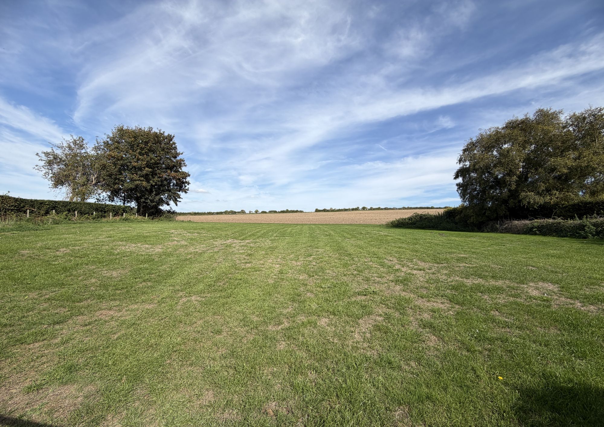 A wide mown grass field edged by hedges and two large trees, opening onto a ploughed field and distant tree line, under a blue sky streaked with high cloud.