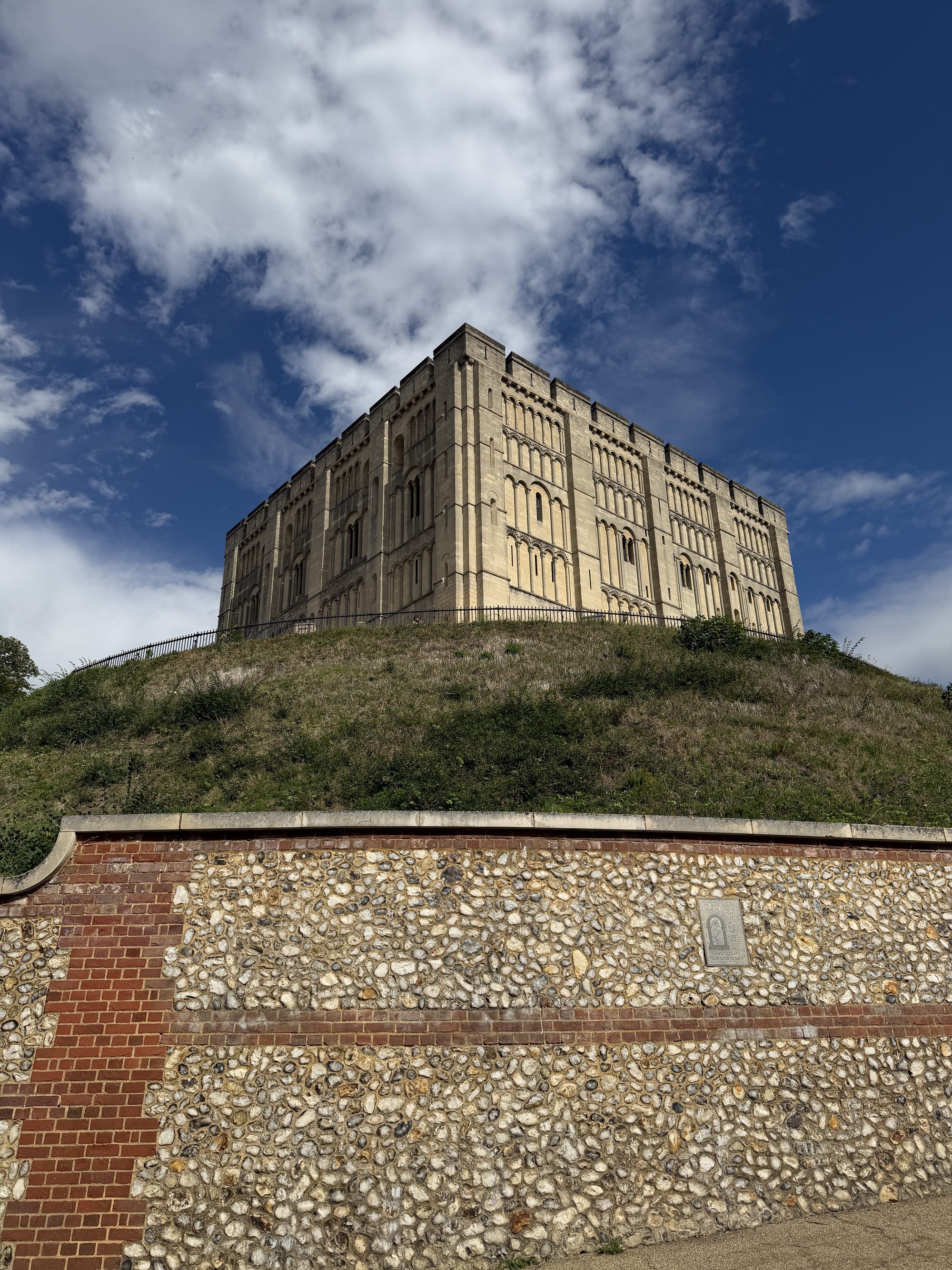 The square stone keep of Norwich Castle on its grassy mound, seen from below beyond a flint and brick wall, under a blue sky with scattered cloud.
