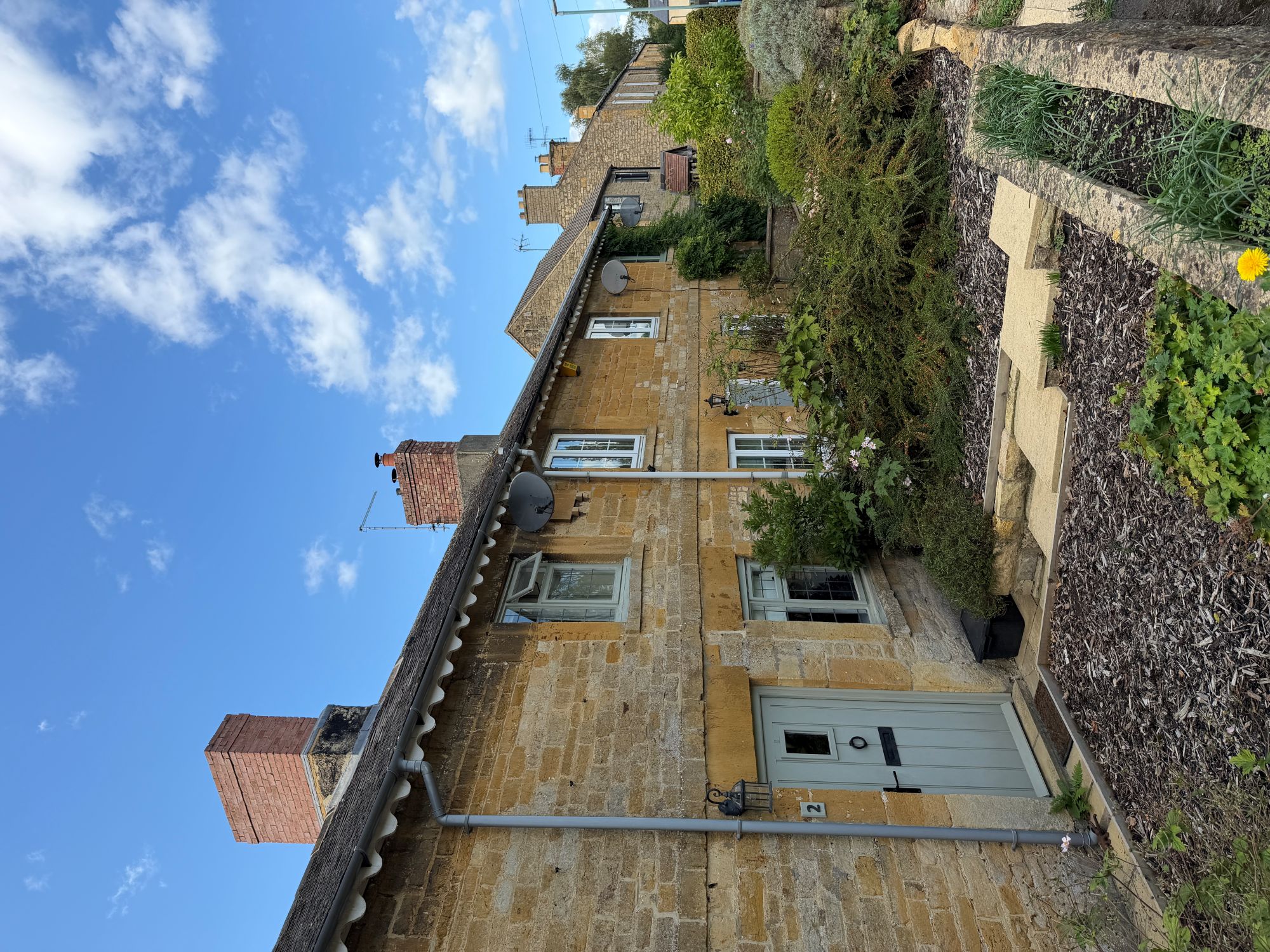 A terrace of honey-coloured stone cottages with brick chimneys and a pale green front door, behind a small garden of shrubs and stone steps, under a blue sky with white clouds.