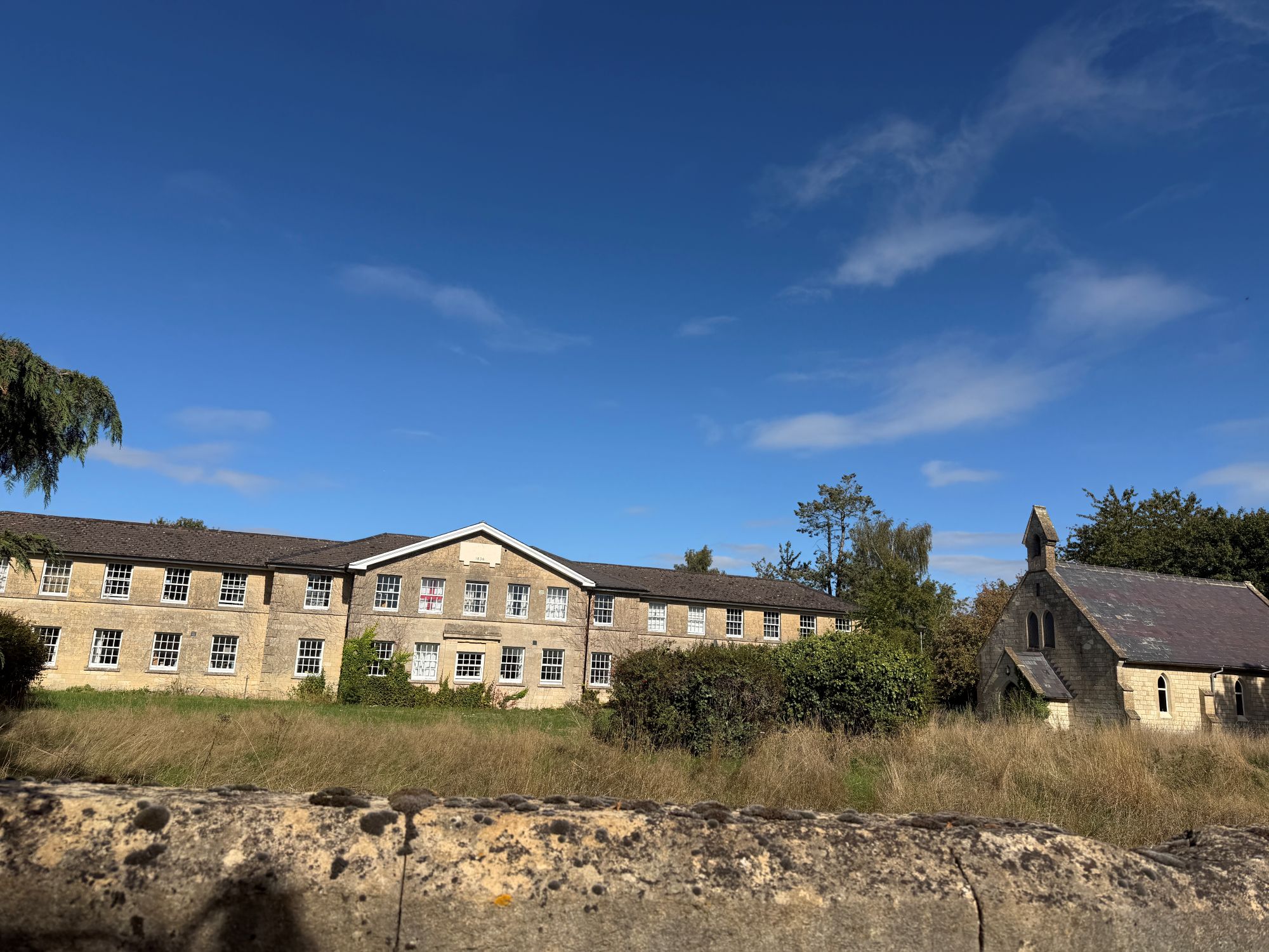 A long two-storey stone building with a central gable and many white-framed windows, behind long grass and a stone wall, with a small stone chapel on the right under a deep blue sky.