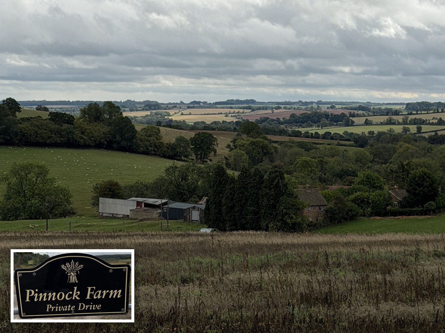 Panoramic view over fields and wooded hills near Didbrook, Gloucestershire, with the buildings of Pinnock Farm in the valley and sheep grazing in the field on the left; in the foreground is an inset of the farm gate sign reading Pinnock Farm, Private Drive