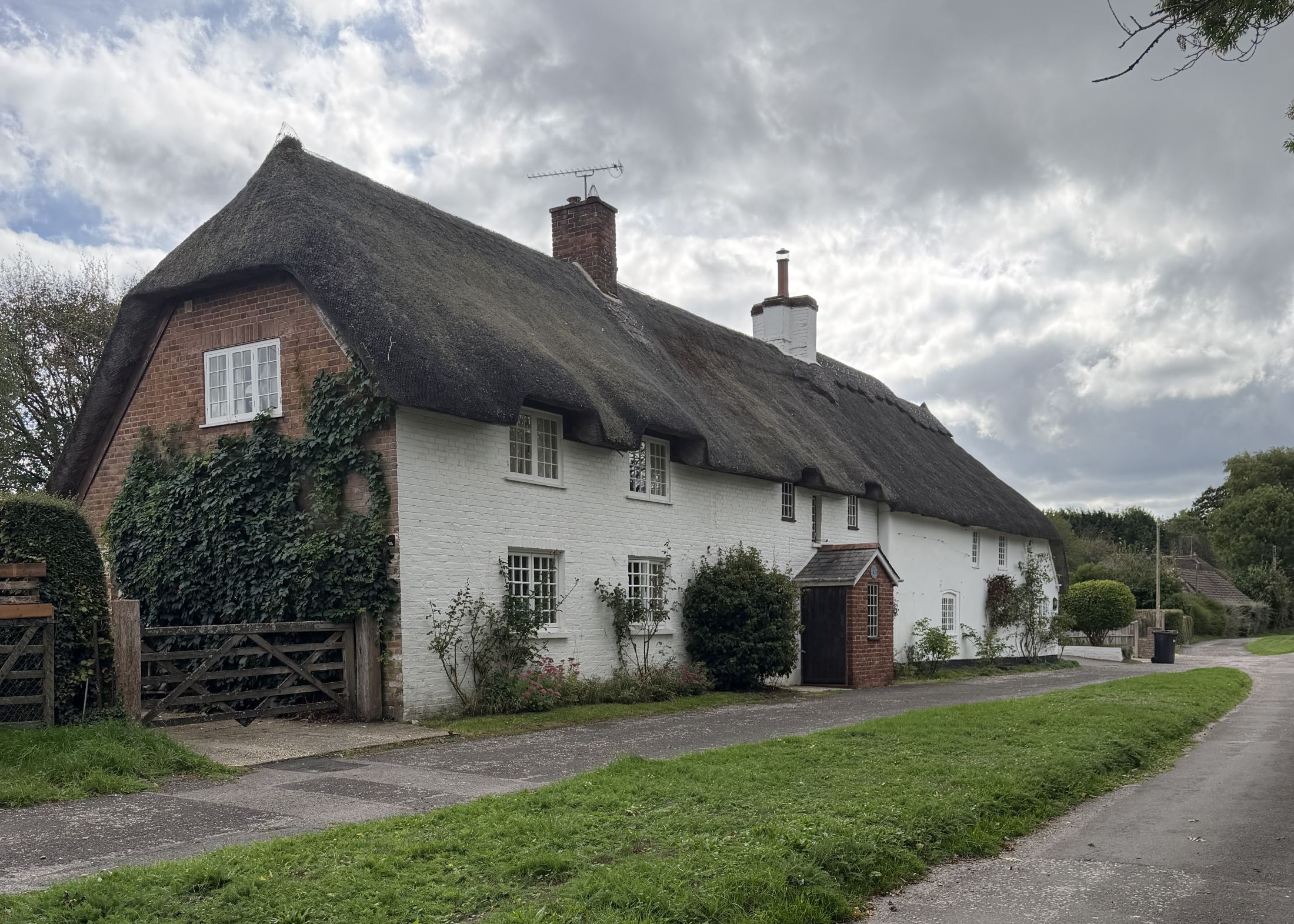 A long white-painted thatched cottage with a red brick gable end covered in ivy, brick chimneys, a small brick porch and a wooden field gate, beside a lane and grass verge under a cloudy sky.