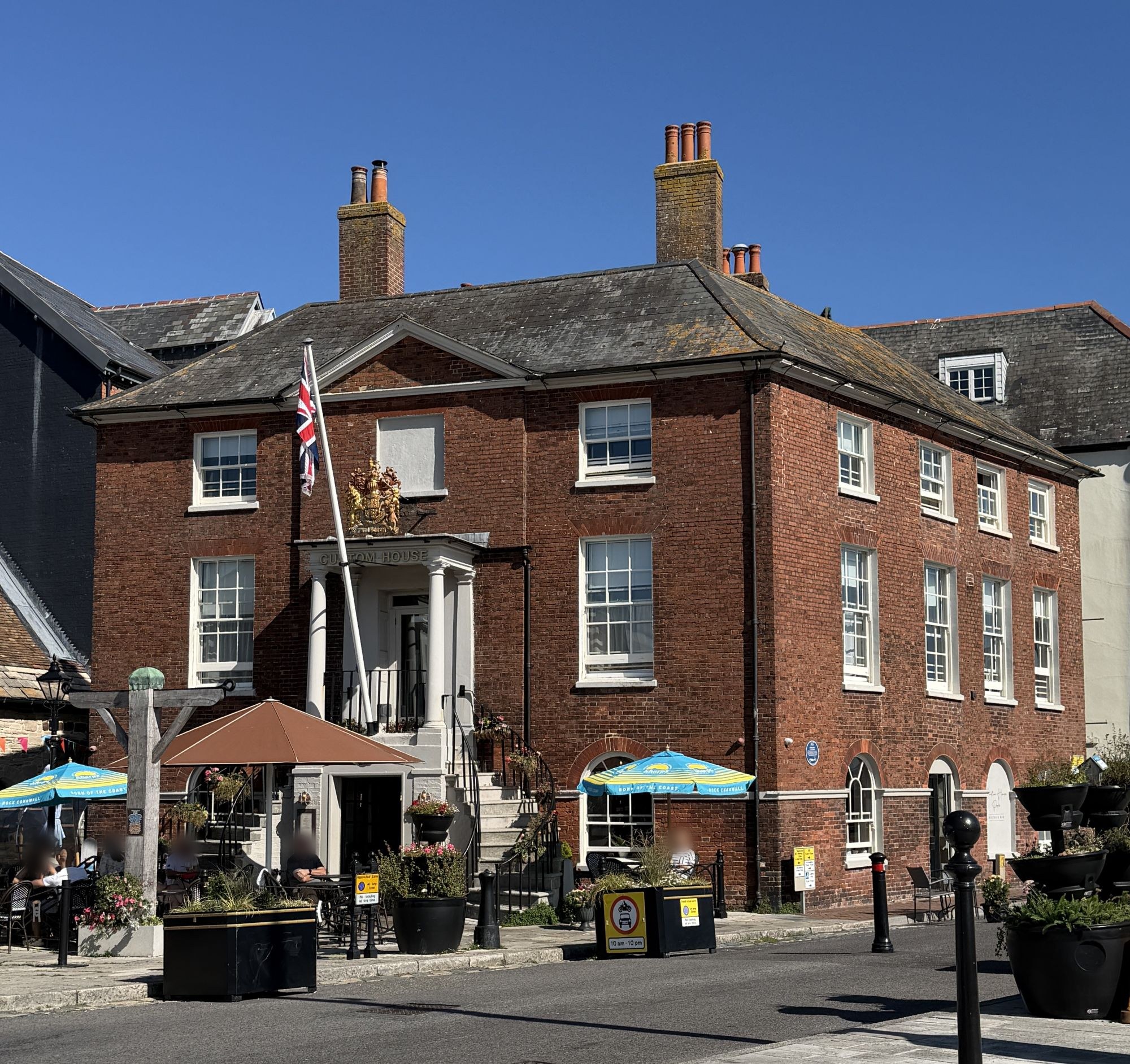 A three-storey red brick Georgian building with a hipped slate roof, tall chimneys and white sash windows, a double segmental staircase rising to a columned porch with a gilded coat of arms and a Union flag above, and the old wooden wool scales in front partly hidden by a caf&eacute; umbrella, with caf&eacute; tables and planters along the quay under a clear blue sky.