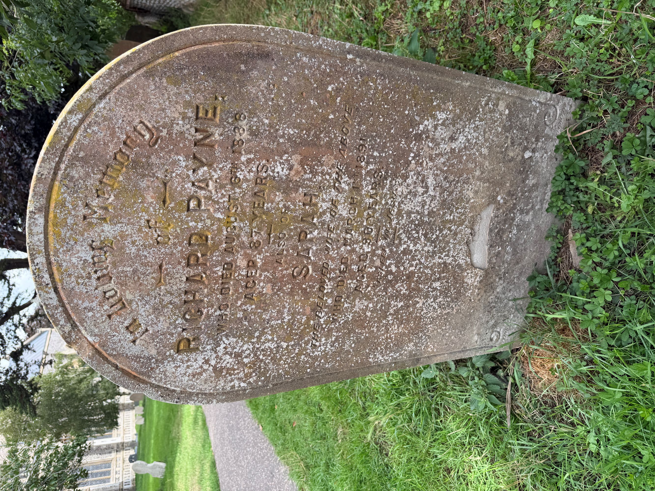 A weathered, lichen-covered arched headstone in grass, inscribed with the names Richard Payne and Sarah, with a path and part of the church behind.