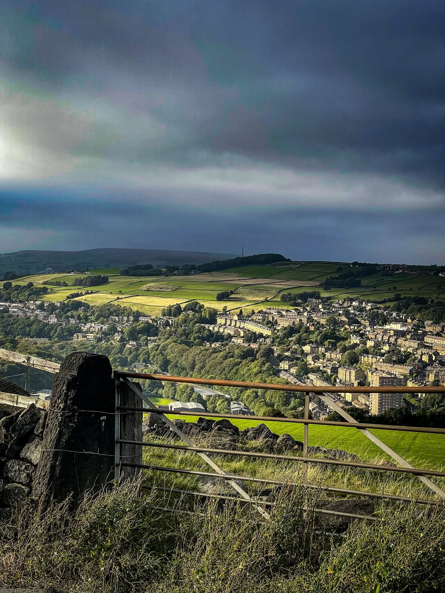 View across a steep green valley to terraced stone houses on the hillside, seen over a metal field gate and stone gatepost, under dark storm clouds with a patch of sunlight on the fields. Photograph by Sean Lewis.