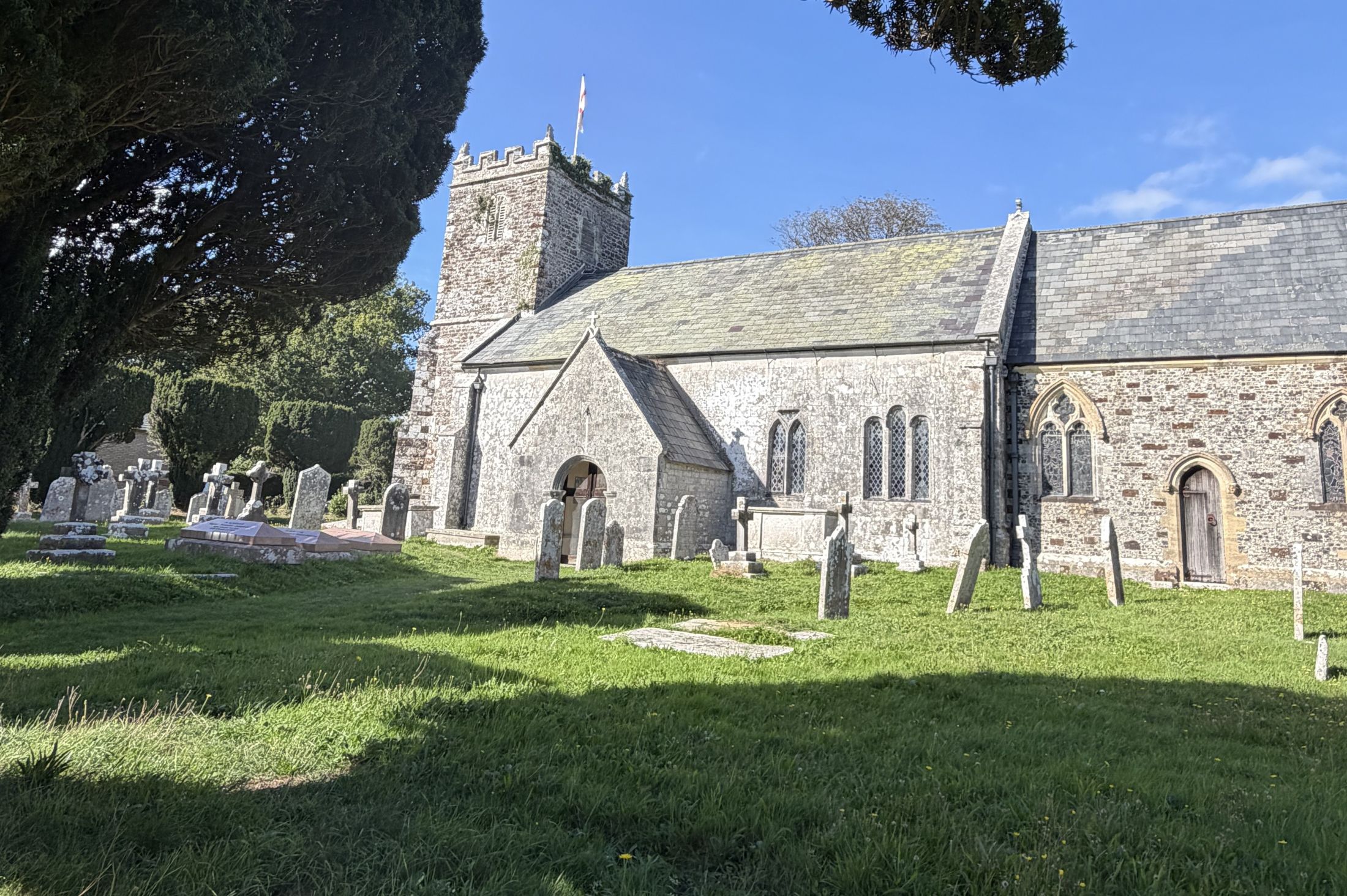 A grey stone church with a battlemented tower flying a flag and a stone-slate roof, seen across a sunlit churchyard of headstones, with the dark branches of a large yew tree on the left under a deep blue sky.