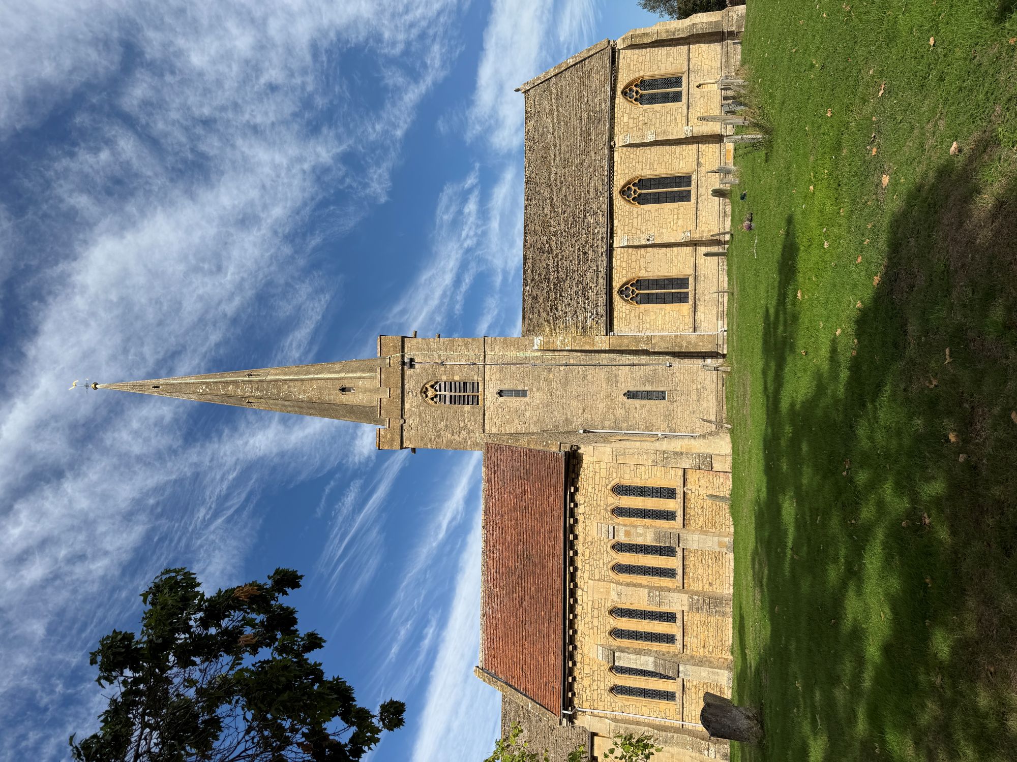 A honey-coloured stone church with a tall tower and slender spire, a red tiled roof on one side, tall pointed windows and old gravestones on a lawn, under a blue sky with wispy cloud.