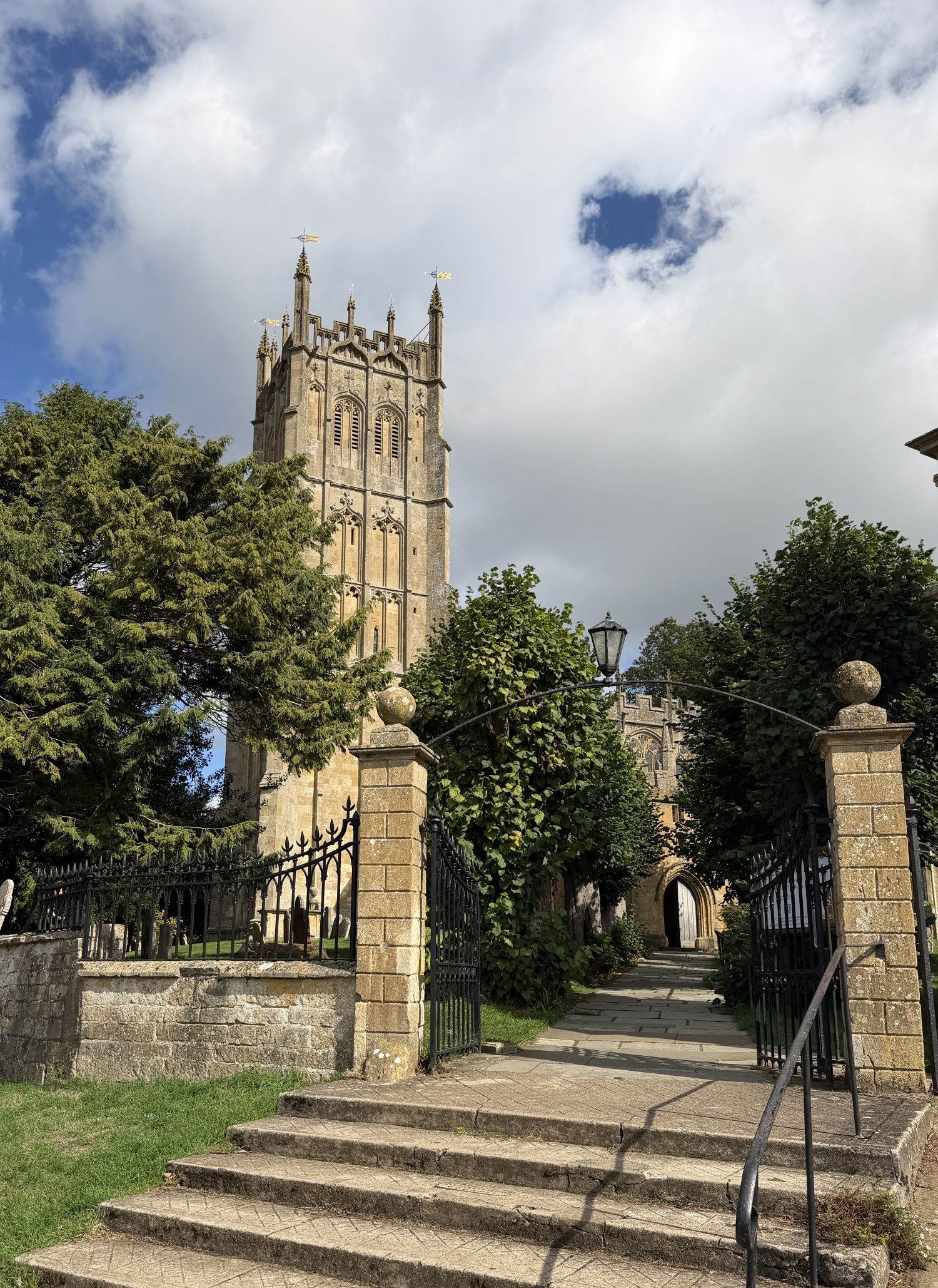 A tall golden stone church tower with pinnacles rises behind stone gate piers, iron gates and leafy trees, above a flight of stone steps and a path, under a cloudy sky.