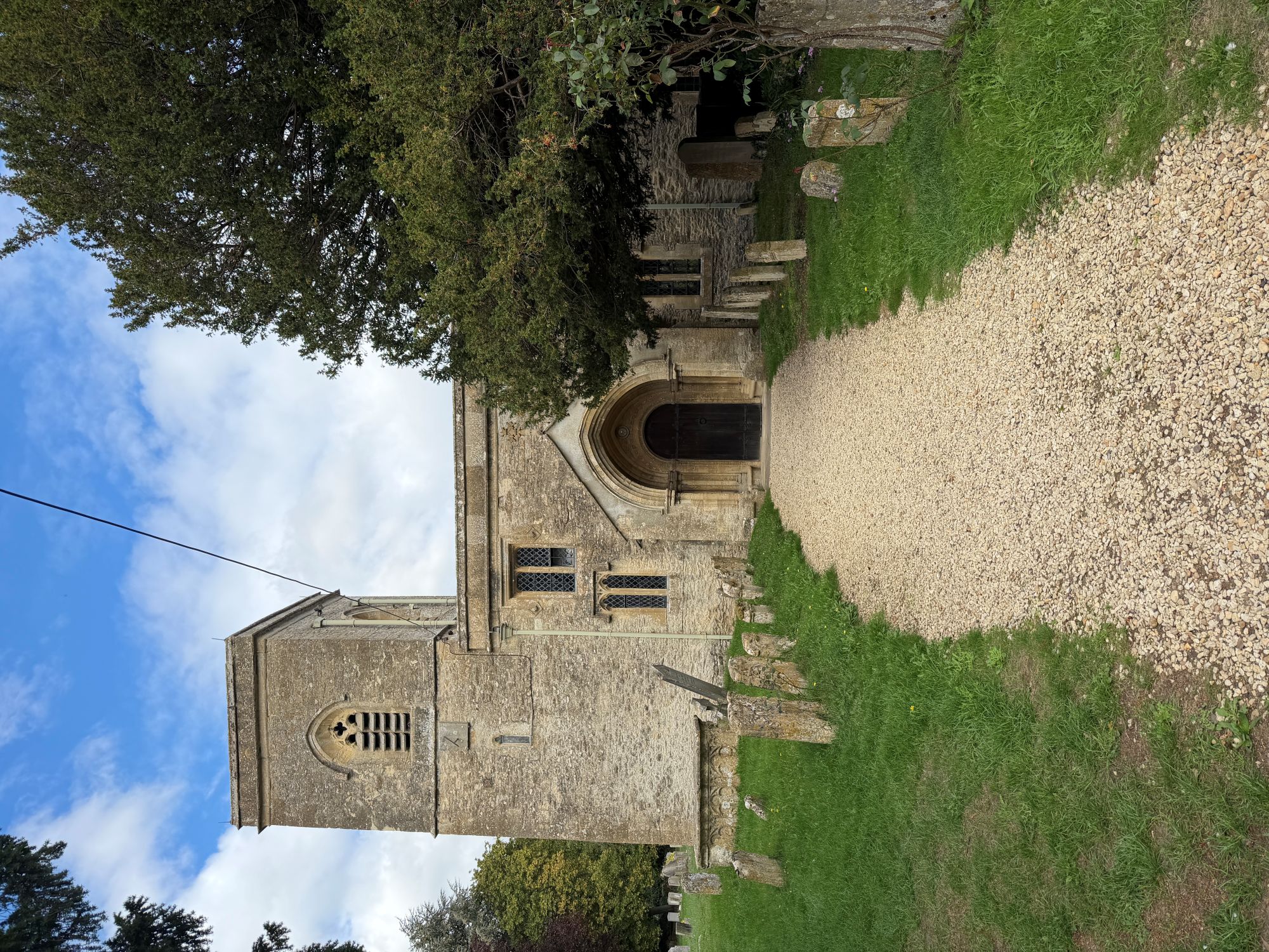 A stone church with a square tower and arched porch stands behind a gravel path, with old headstones on the grass and a large dark yew tree at right, under a blue sky with white clouds.
