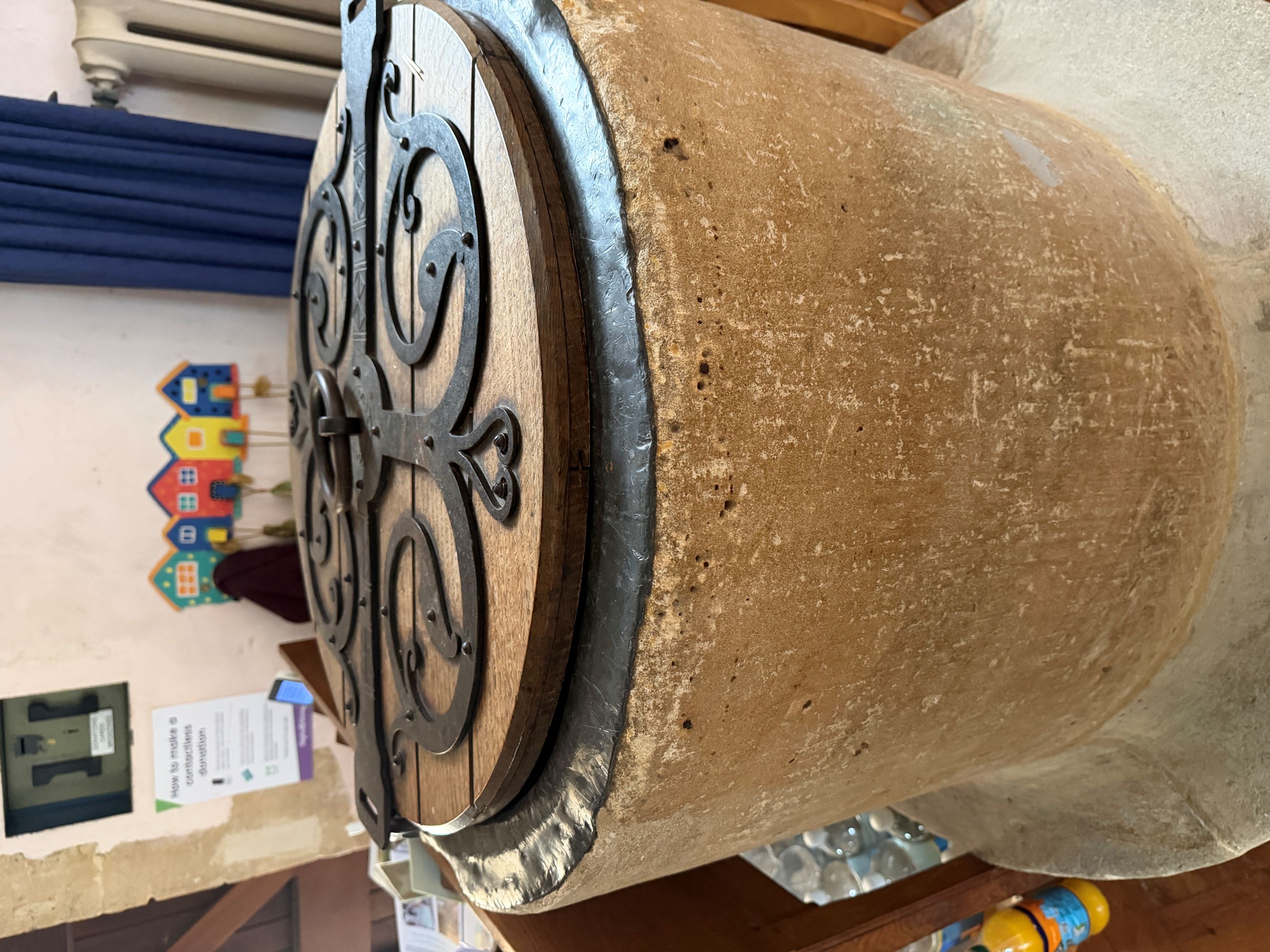 A round stone font with a dark wooden lid and decorative scrolled ironwork, standing in a church corner with a blue curtain and small colourful paper houses on the wall behind.