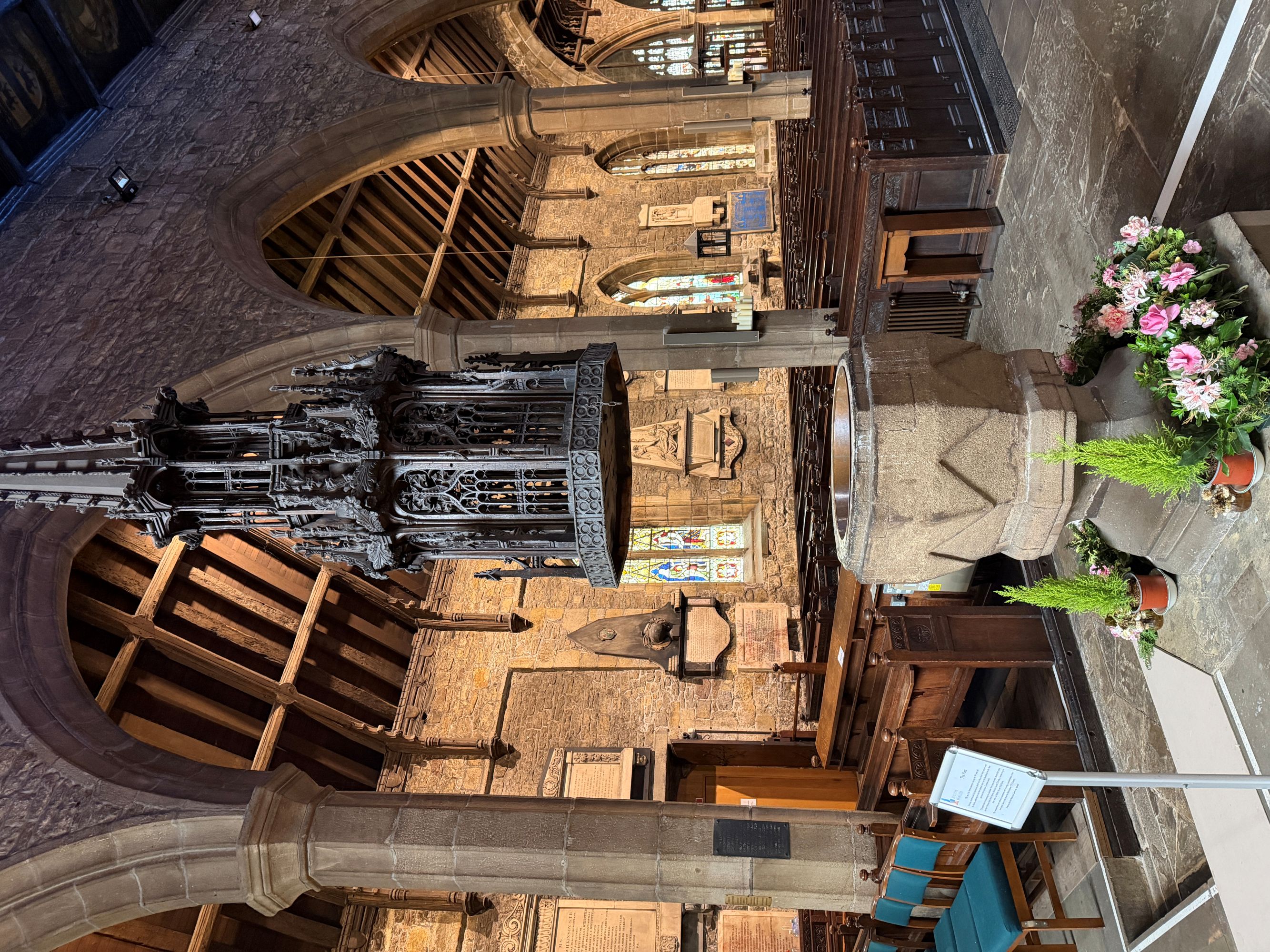A carved stone font with flowers around its base, under a tall dark carved wooden spire-shaped cover hanging from above, in a church with oak pews and a timber roof.