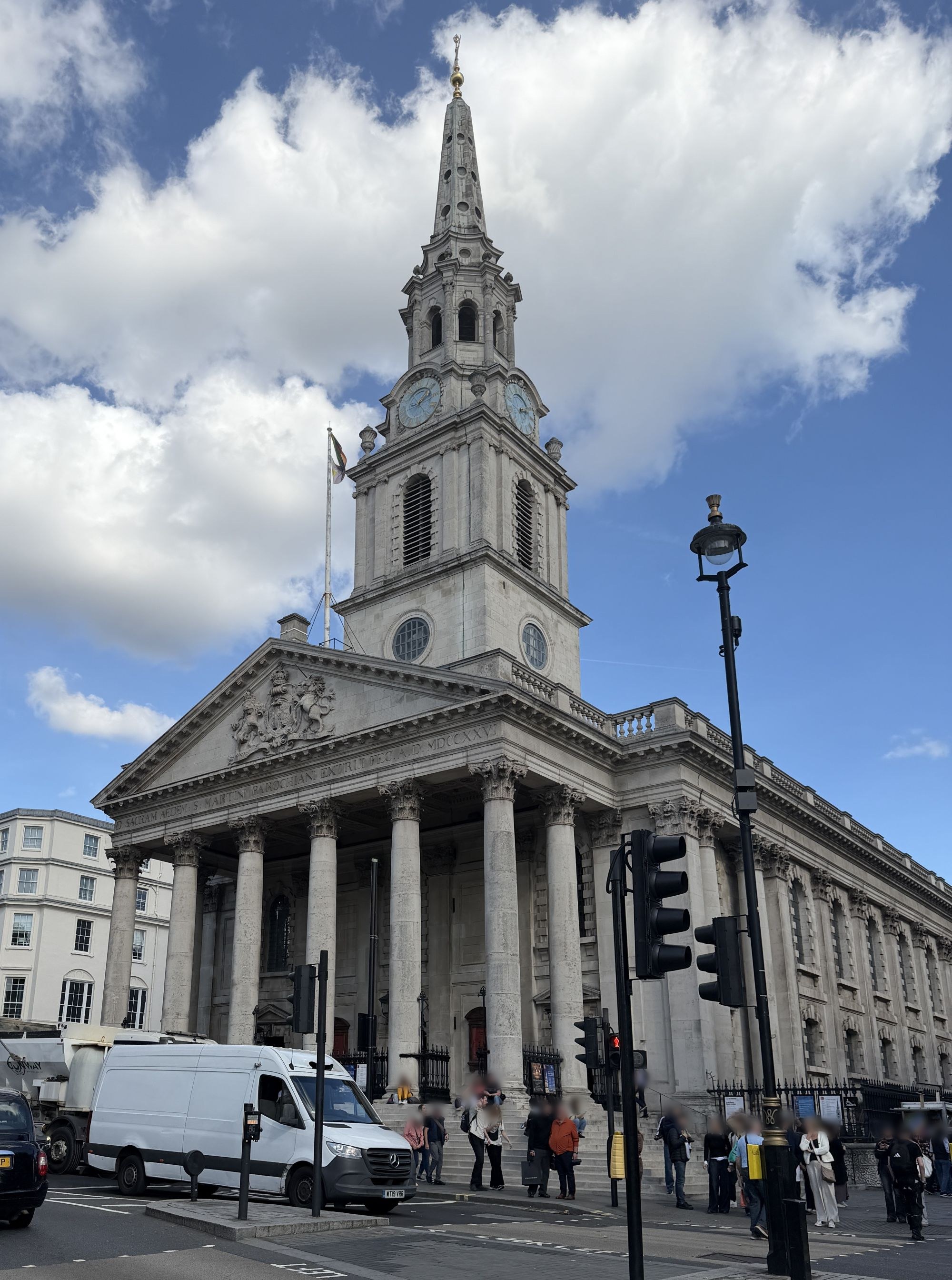 A stone church with a tall columned front and a clock tower and spire rises above a busy city street with traffic lights, a white van and people on the steps, under a blue sky with white clouds.