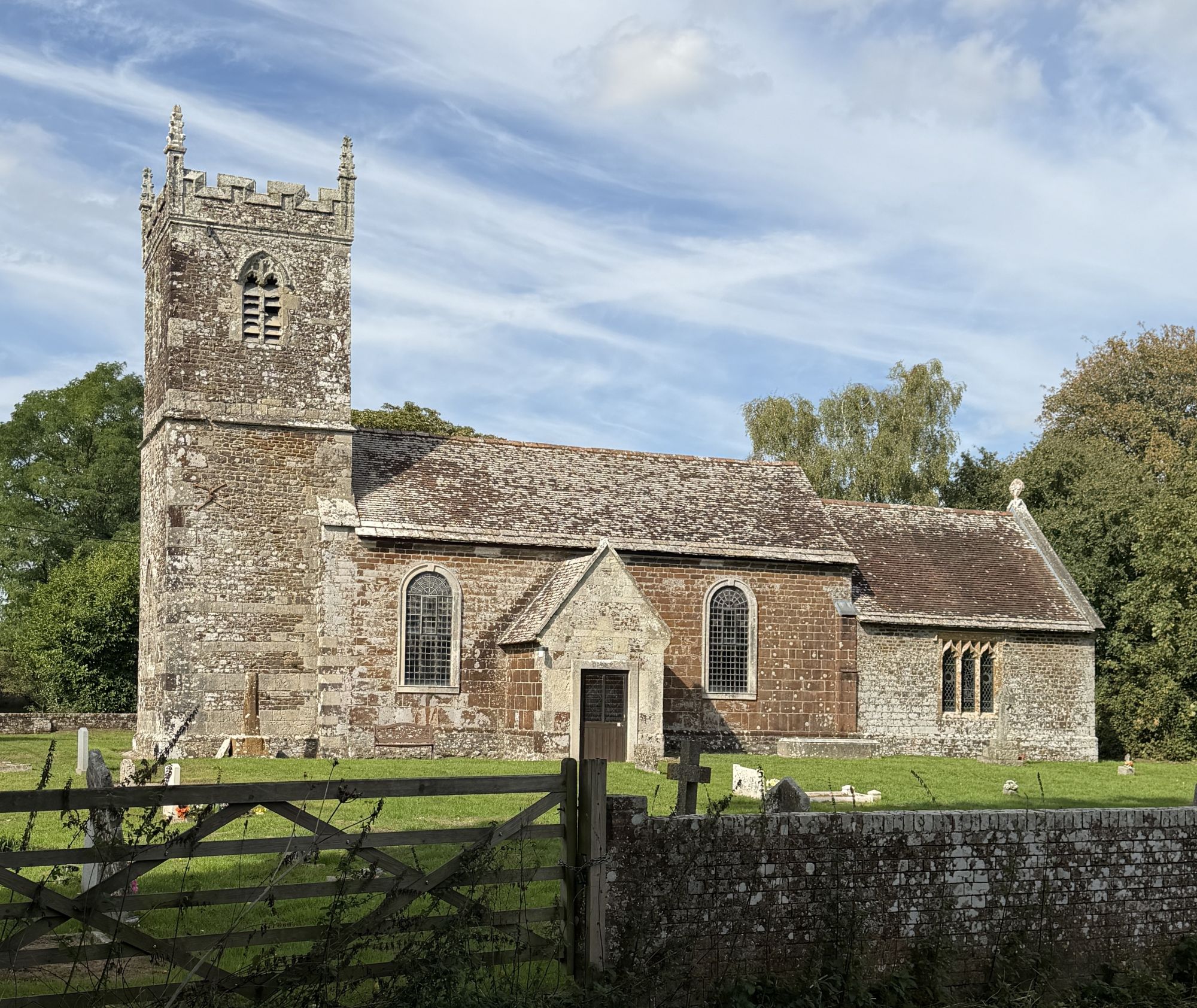 A small stone church with a tall square tower topped by battlements and pinnacles, a stone porch and round-headed windows, seen over a stone wall and wooden field gate, under a blue sky with thin cloud.