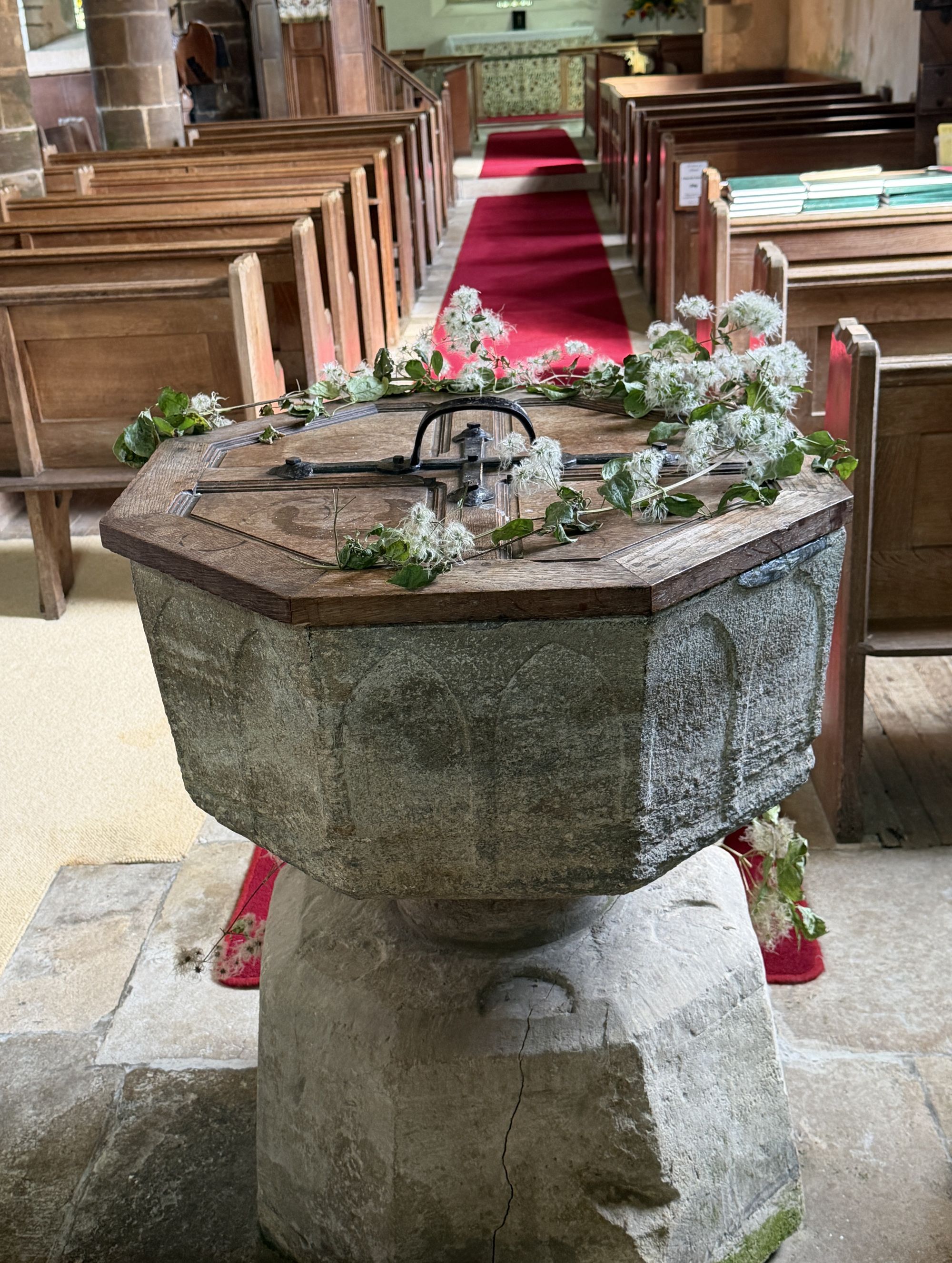 A plain octagonal stone font with a carved arched pattern and a wooden lid with an iron handle, decorated with sprigs of green leaves and white fluffy flowers, in front of wooden pews and a red-carpeted aisle.