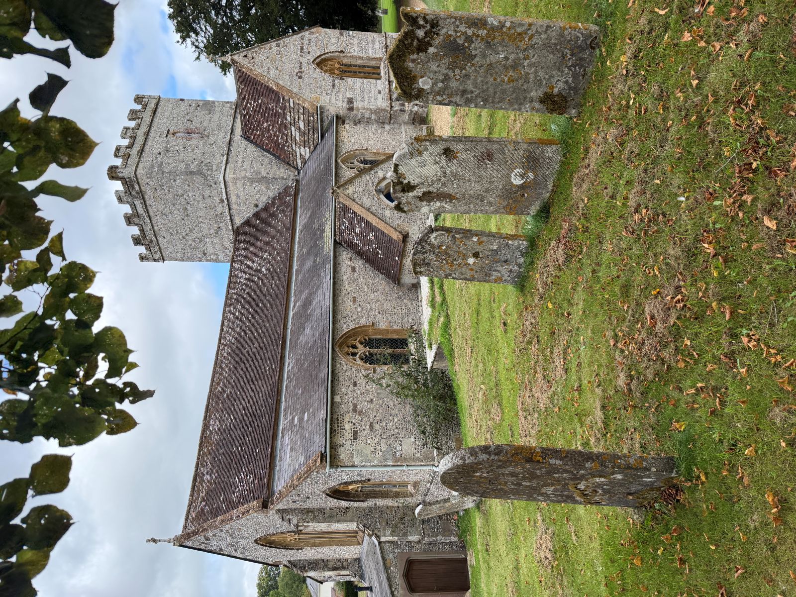 A flint and stone church with a battlemented tower, slate roofs and pointed windows, seen across a grassy churchyard with weathered headstones, under overhanging leaves and a cloudy sky.