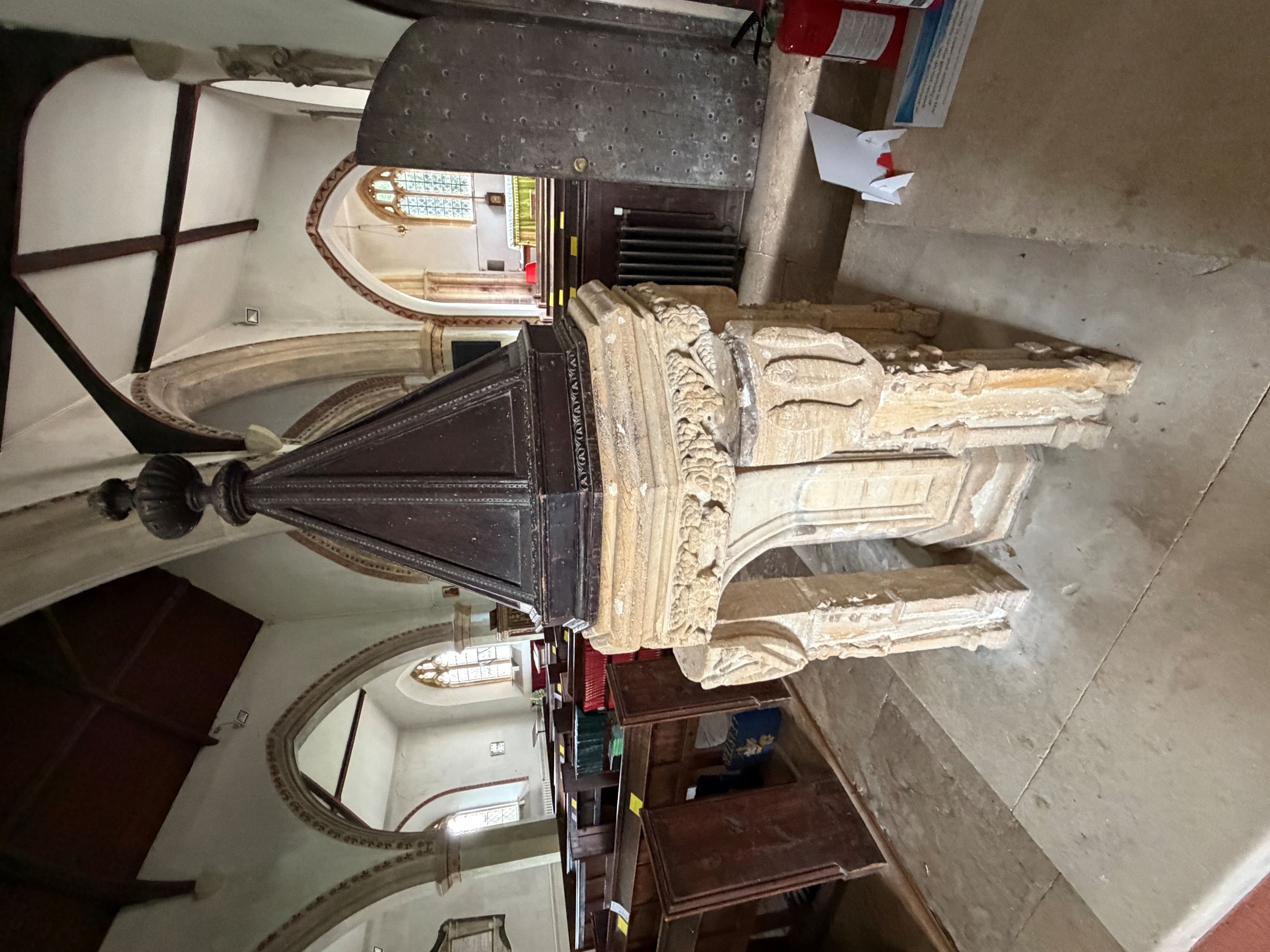 A carved pale stone font with a tall dark pointed wooden cover topped by a finial, inside a church with pointed stone arches, wooden pews and a stained glass window beyond.