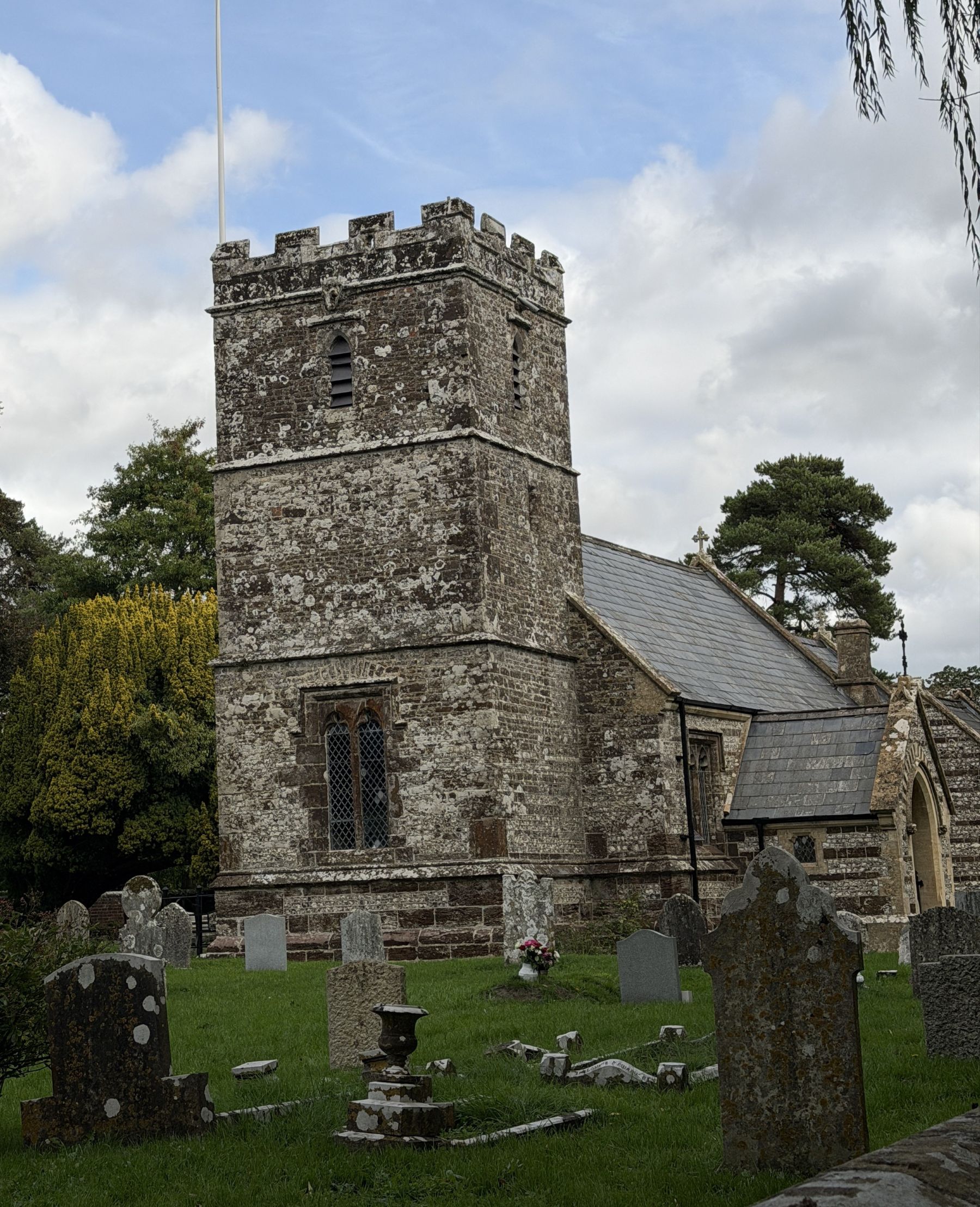 A lichen-covered stone church tower with battlements and a leaded window, beside a slate-roofed nave, seen across a churchyard crowded with old headstones, under a cloudy sky.