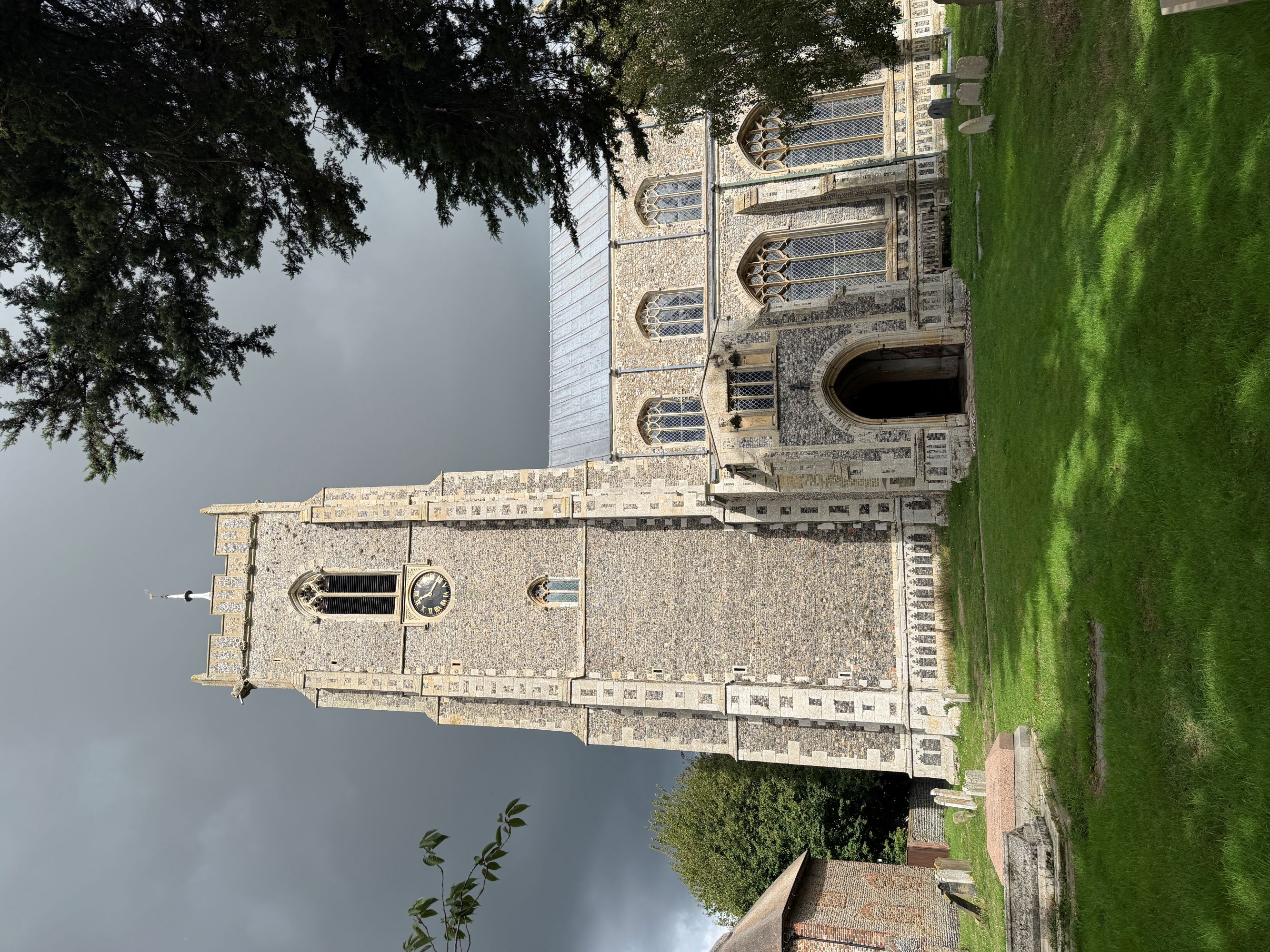 The flint and stone tower and porch of a large church seen across a graveyard lawn, with dark storm clouds behind.