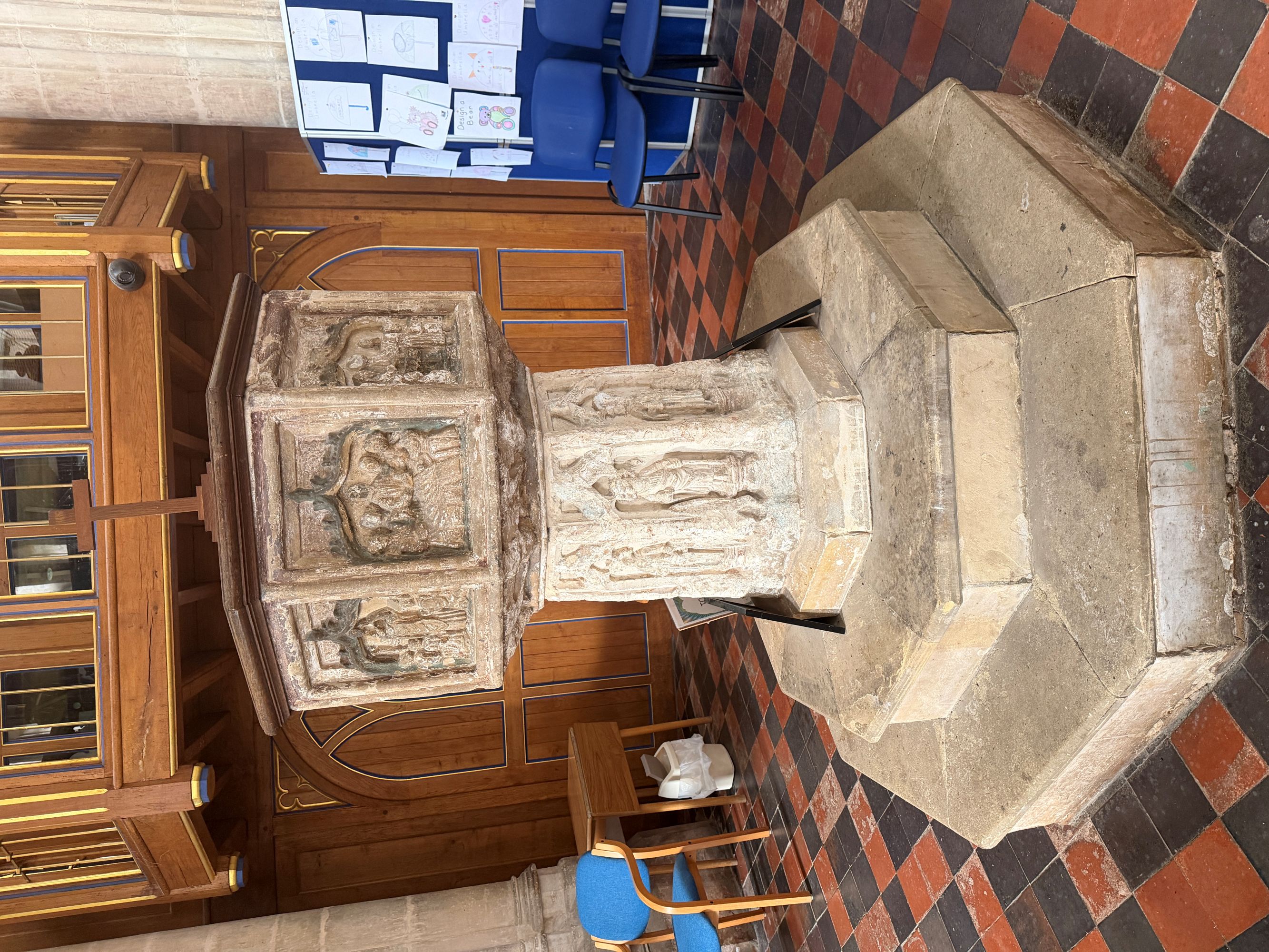 A carved stone octagonal baptismal font with figures on its bowl and stem, standing on a stepped base on a tiled church floor.
