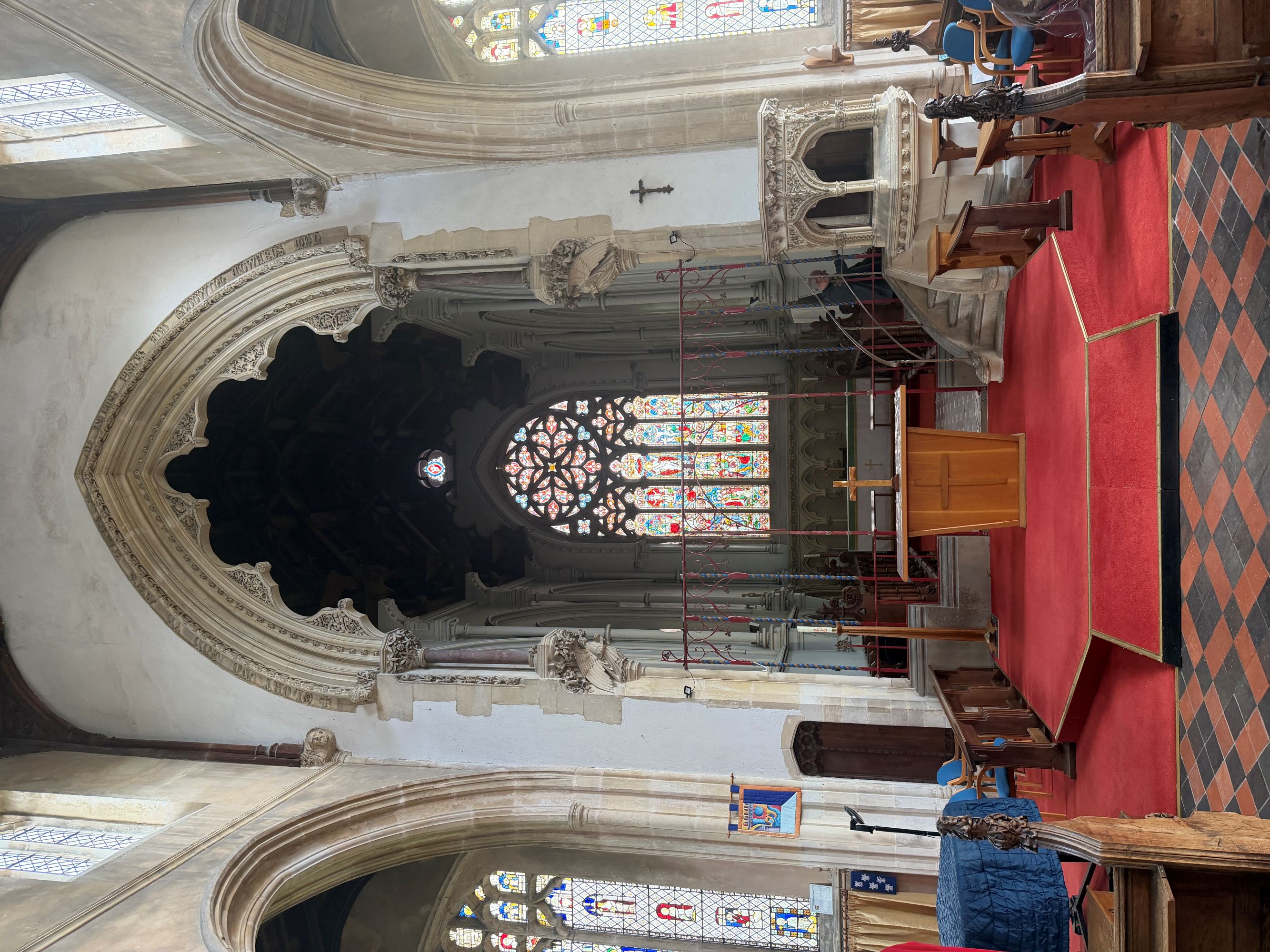 The interior of a church looking towards the chancel, with a carved stone arch, a large stained glass window above the altar, a pulpit on the right and a red carpeted floor.