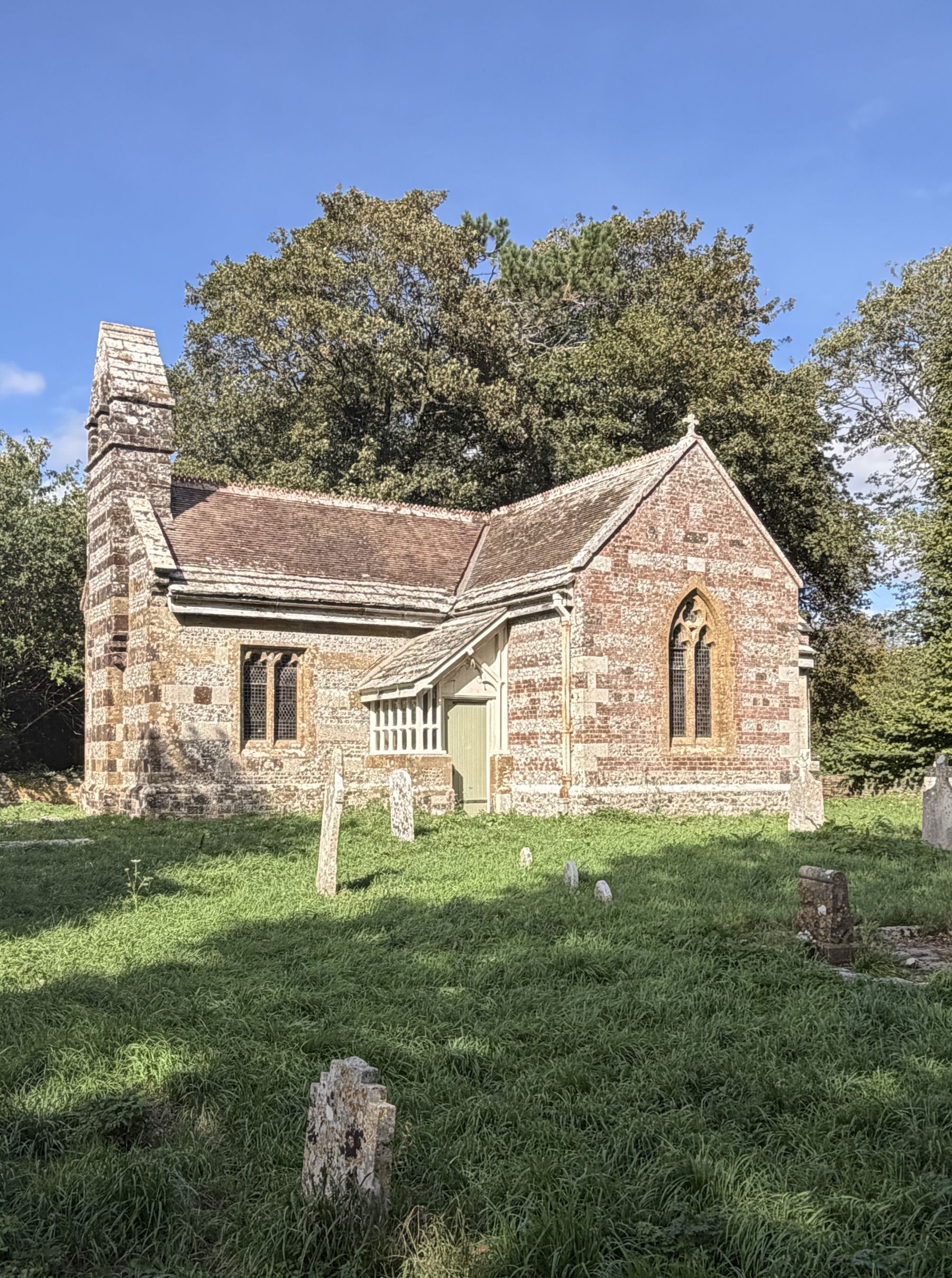 A small stone and brick banded church with a tiled roof, a tall narrow stone stack at one end, a pointed window and a small timber porch, standing in long grass with scattered headstones and a large tree behind, under a blue sky.