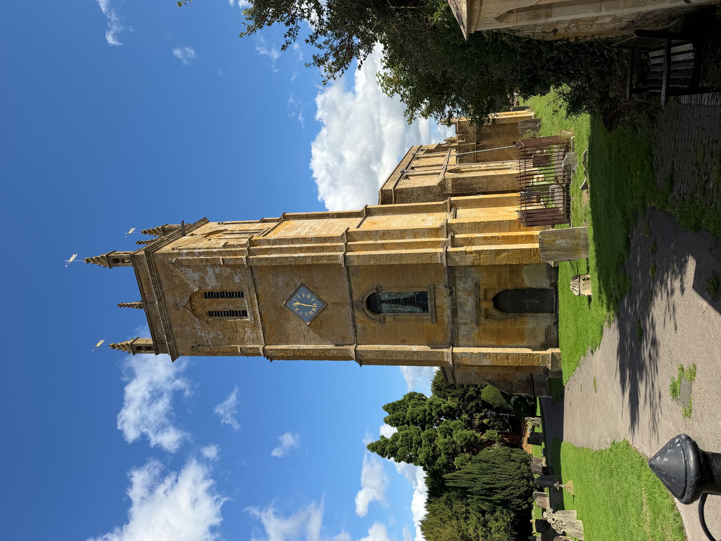 A golden stone church tower with pinnacles and a clock face rises above a grassy churchyard with old gravestones, under a blue sky with white clouds.