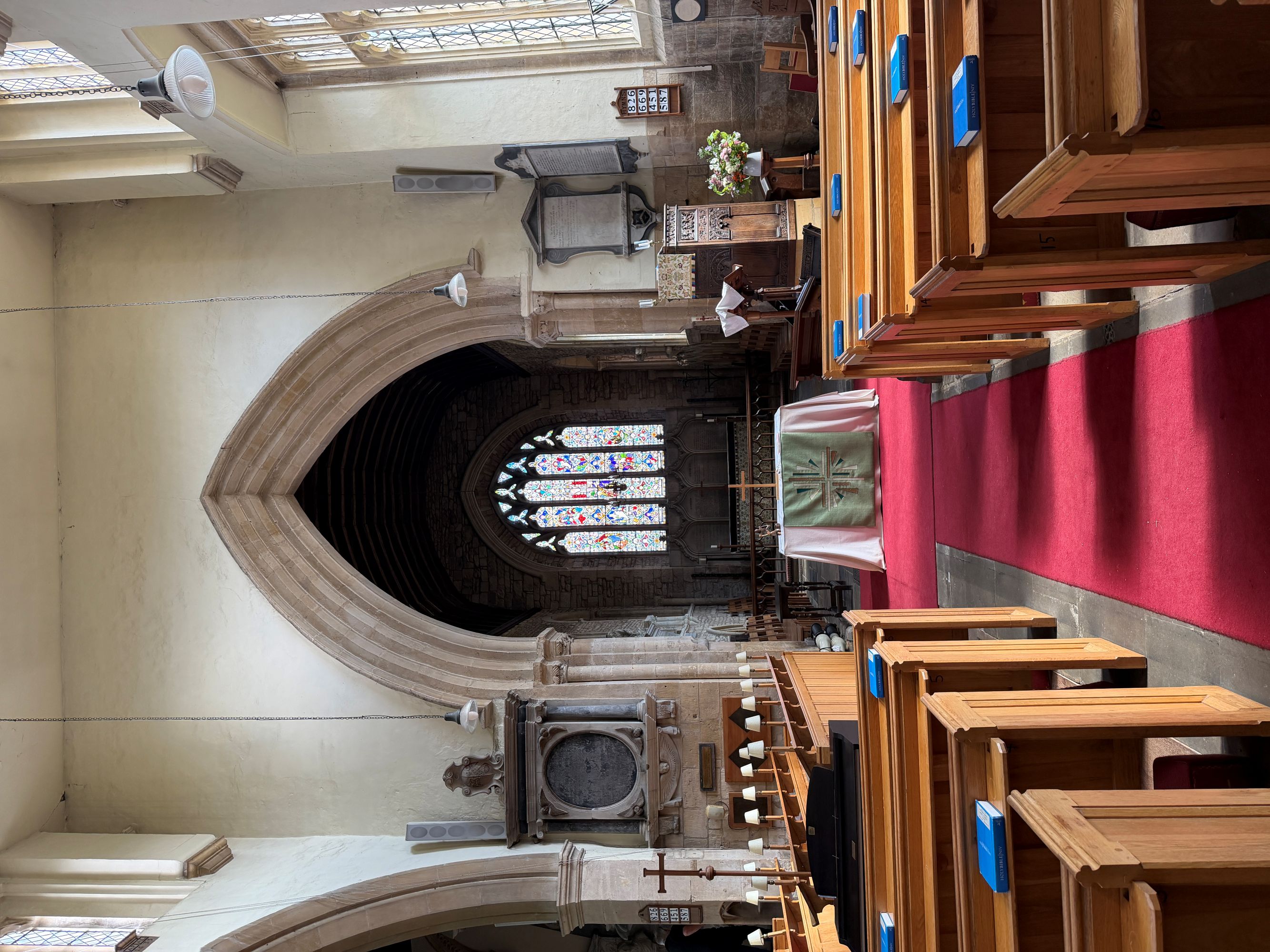A church interior looking down a red-carpeted aisle between wooden pews to an altar and a stained glass window beneath a pointed stone arch.