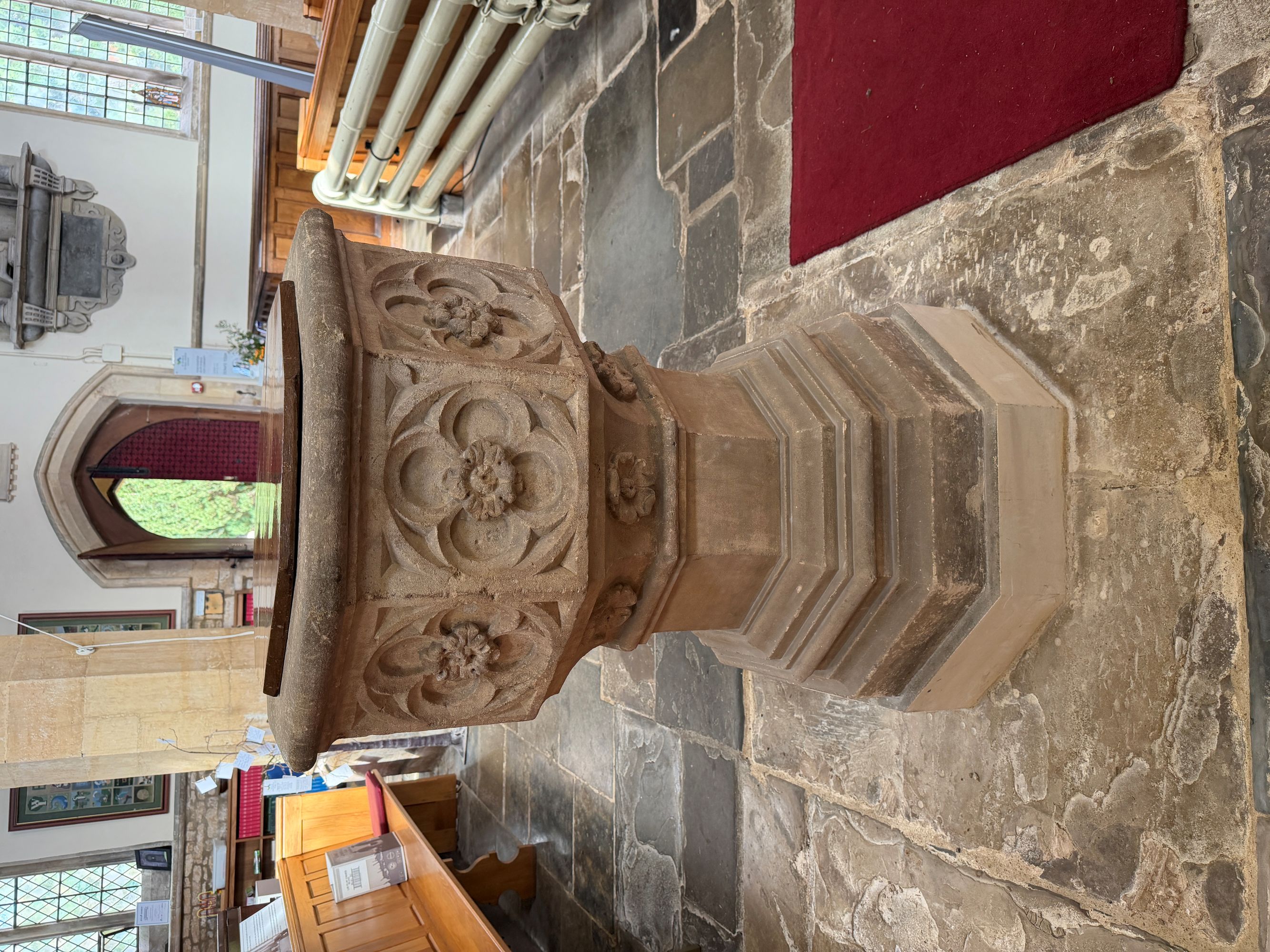 A carved octagonal stone font with flower designs on its sides and a wooden lid, standing on a flagstone floor inside a church with wooden pews and an open arched doorway.