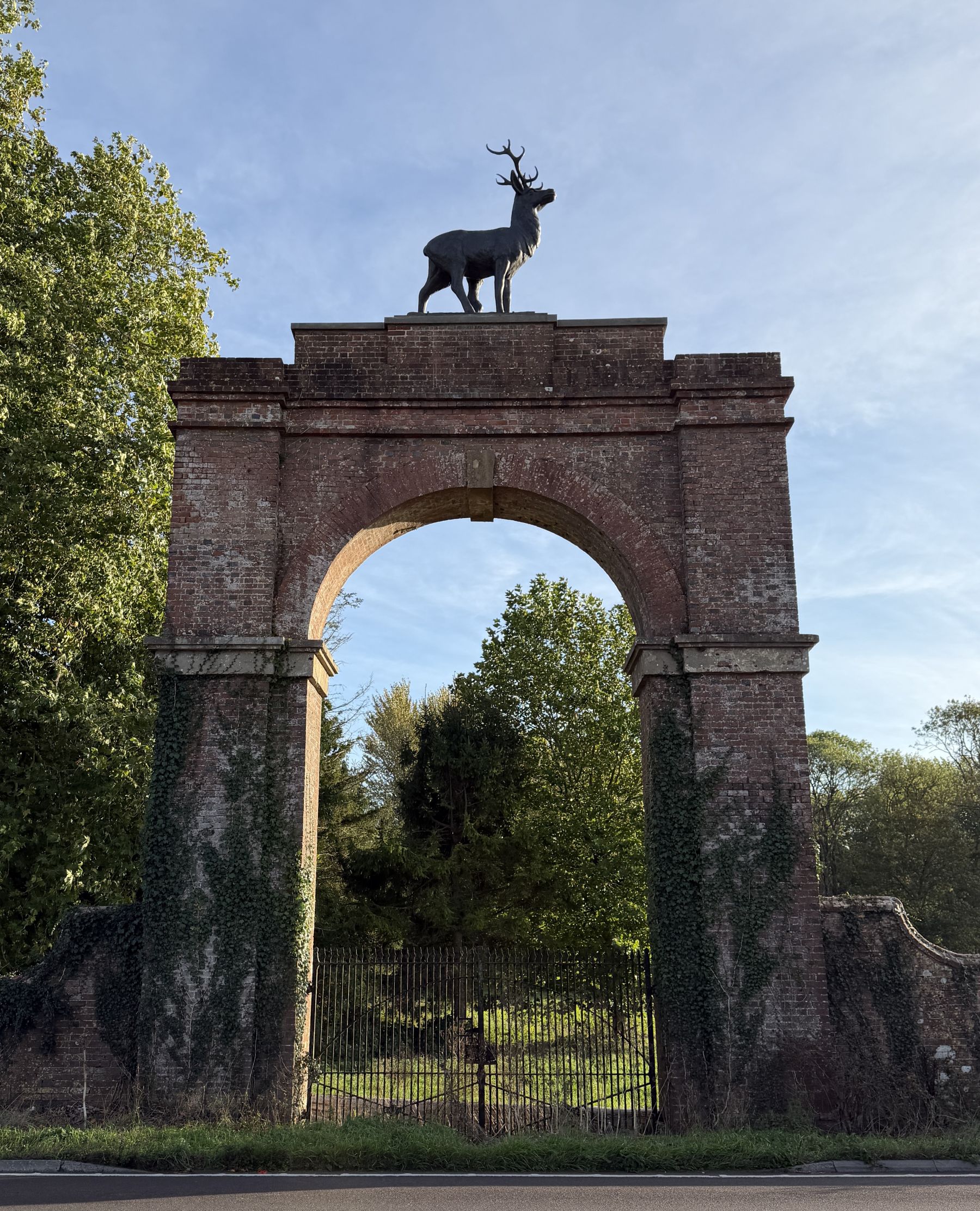 A tall red brick archway with stone bands, topped by a dark statue of a stag with antlers, framing iron railings and trees behind, with ivy on its piers, beside a road under a pale blue sky.