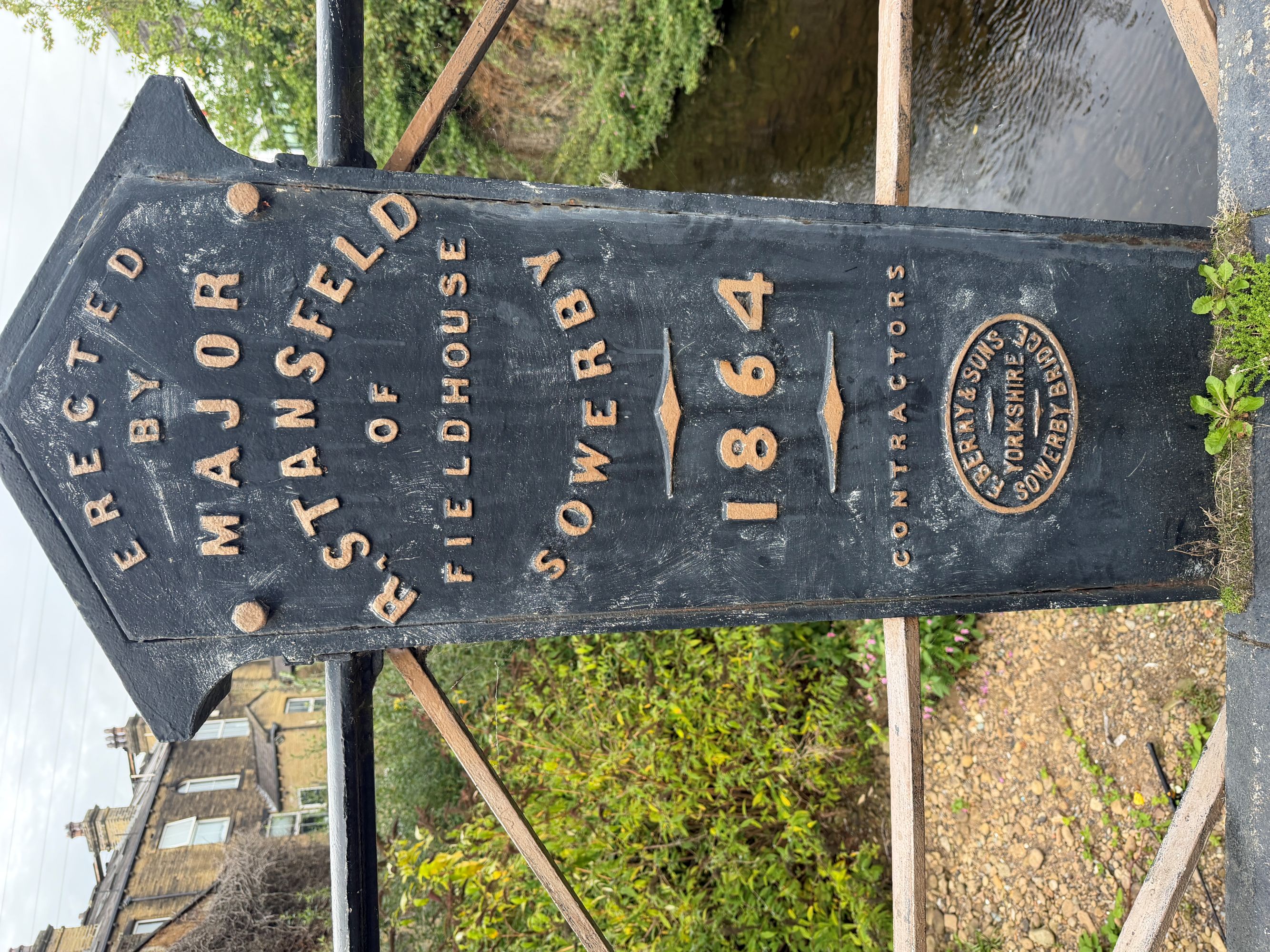 A cast-iron bridge post with a raised plate reading &ldquo;Erected by Major R. Stansfeld of Fieldhouse, Sowerby, 1864, Contractors&rdquo;, above an oval maker&rsquo;s plate reading &ldquo;F. Berry & Sons, Yorkshire, Sowerby Bridge&rdquo;. A river and stone houses are behind.