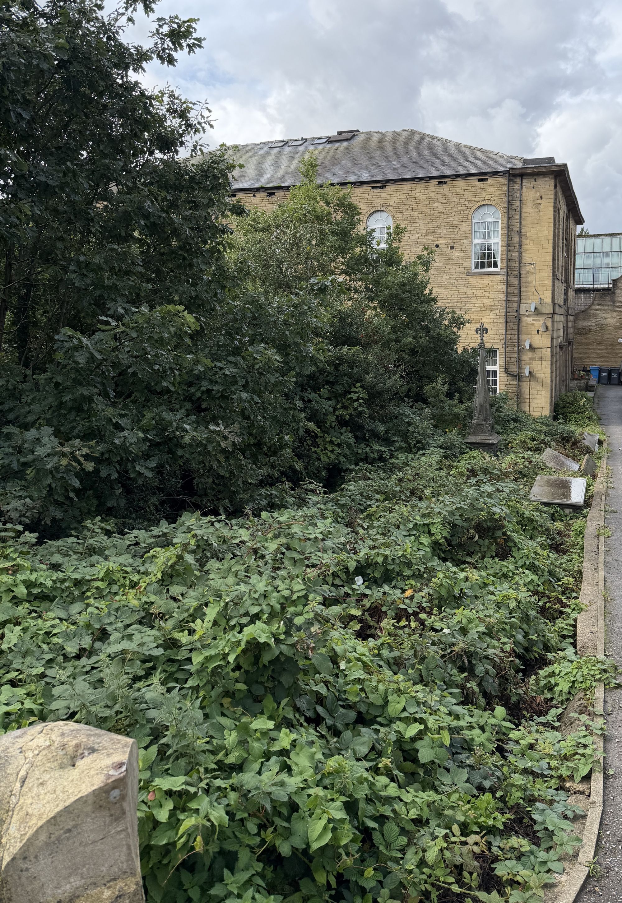 A cemetery overgrown with trees and brambles, with a spire-topped iron monument and flat stone slabs beside a path, and a stone chapel building behind.