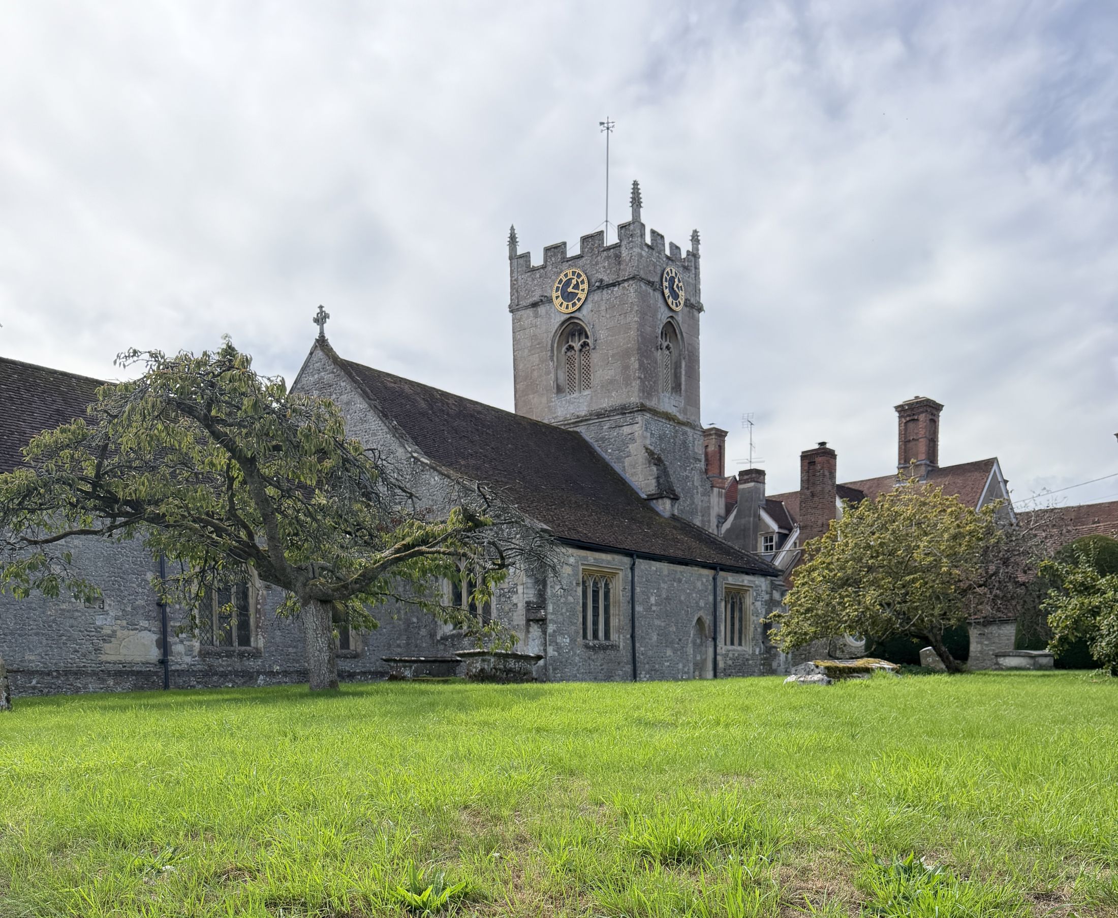A grey stone church with a battlemented clock tower and a dark tiled roof, seen across a lawn with two trees, and red brick chimneys of a large house behind.
