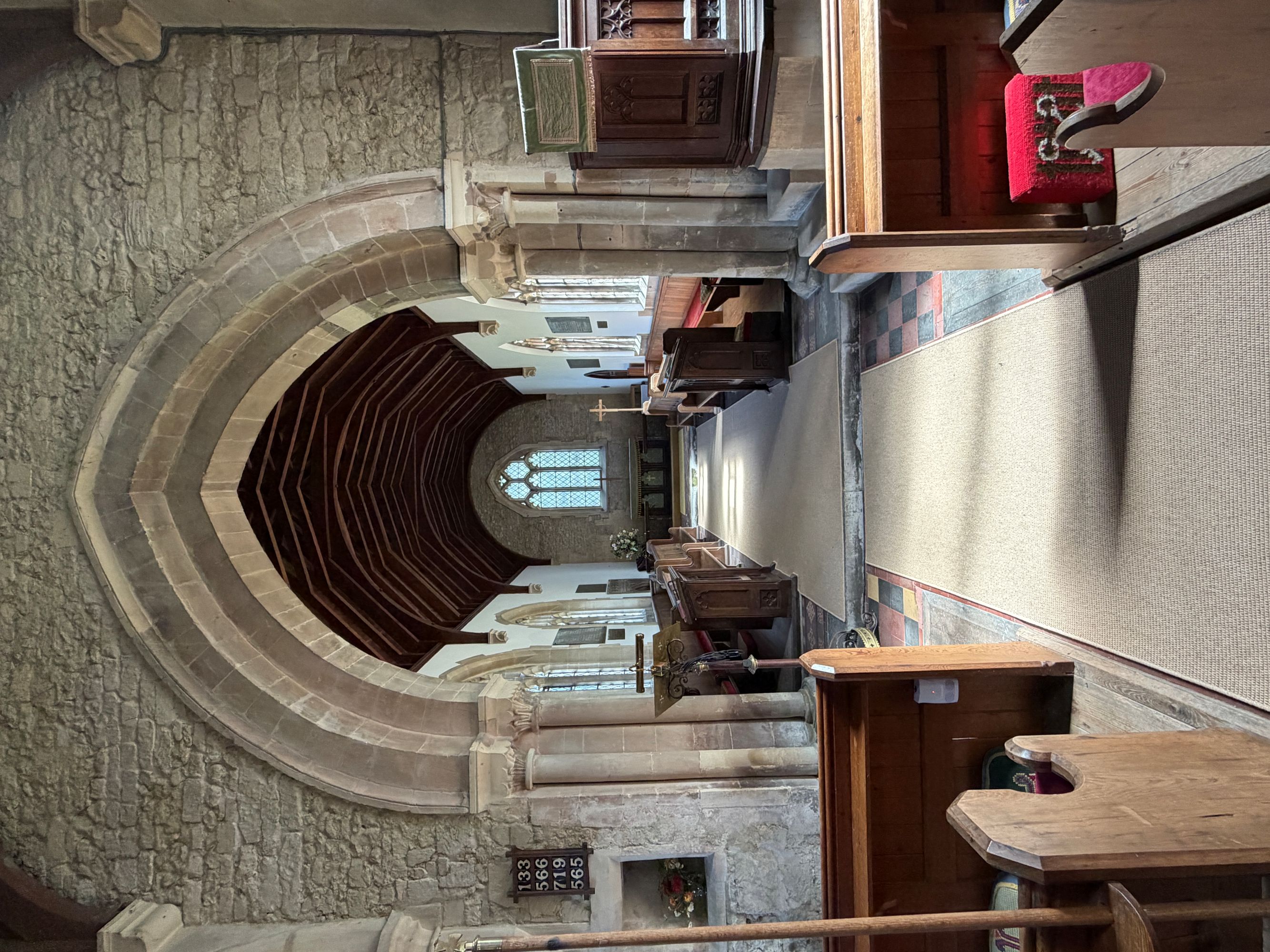 Looking along a church interior through a pointed stone chancel arch to a distant altar and window, with wooden pews, a pulpit on the right and a carpeted aisle.
