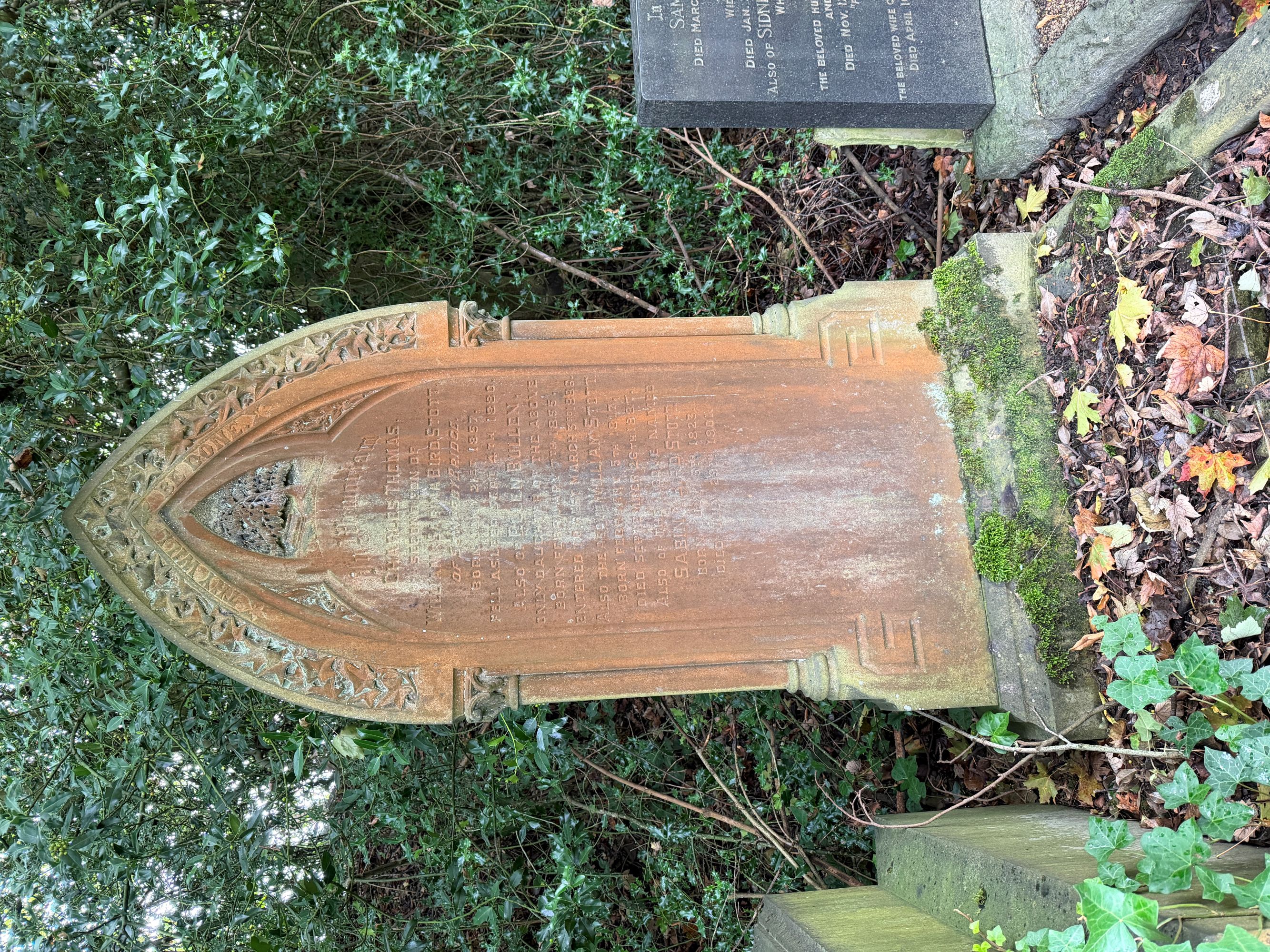 A pointed-arch sandstone headstone with an ivy-leaf border and a weathered inscription, with moss on its base and fallen leaves in front.