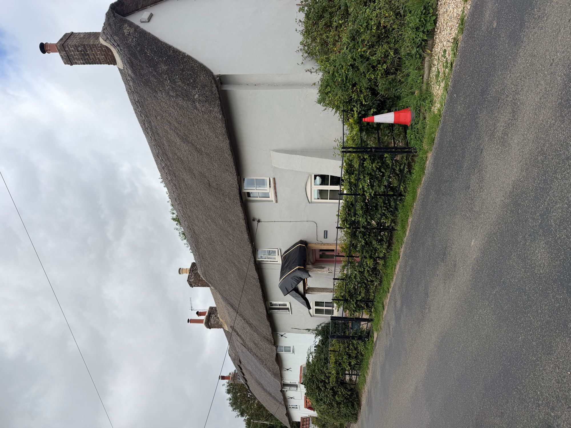 A long white-painted thatched cottage row with brick chimneys, a small porch and low iron railings behind a hedge, beside a quiet village road under a grey sky.